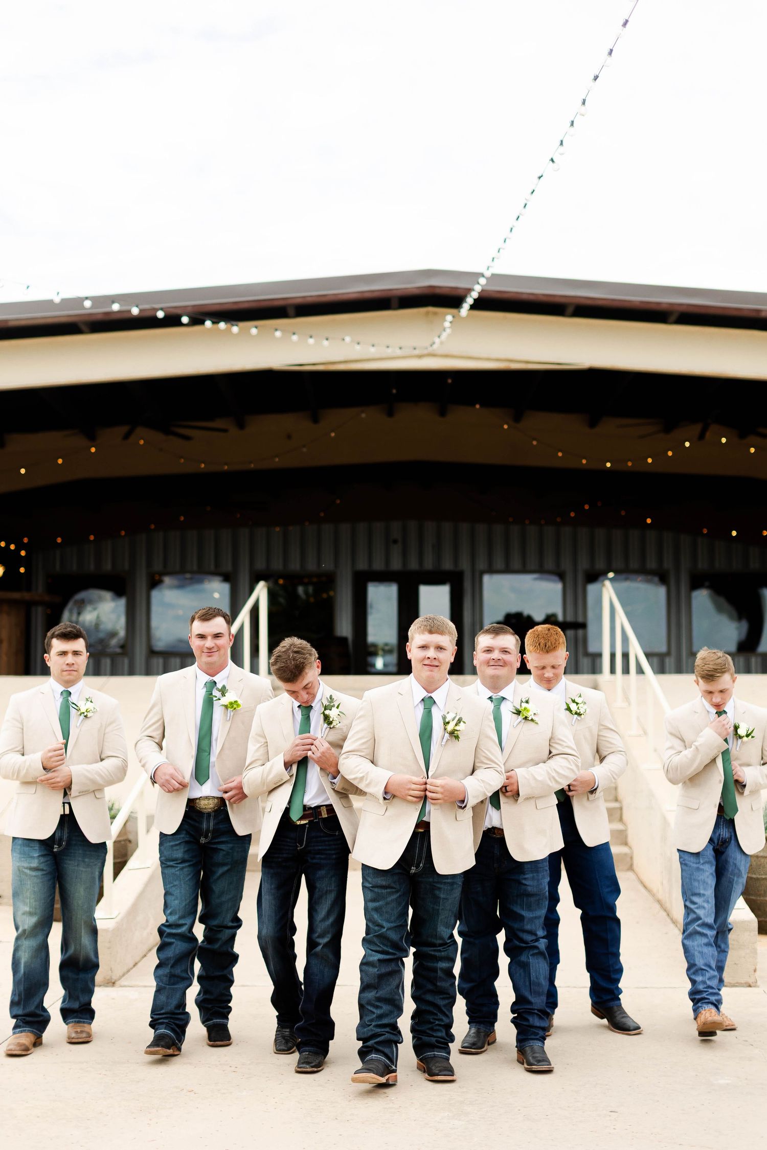 Wedding party in beige jackets and jeans lined up against a rustic venue with string lights.