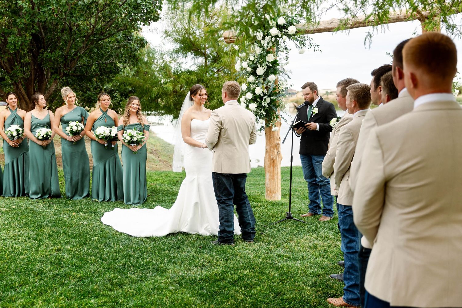 Wedding ceremony scene with rustic wooden arch and guests gathered on grassy lawn.
