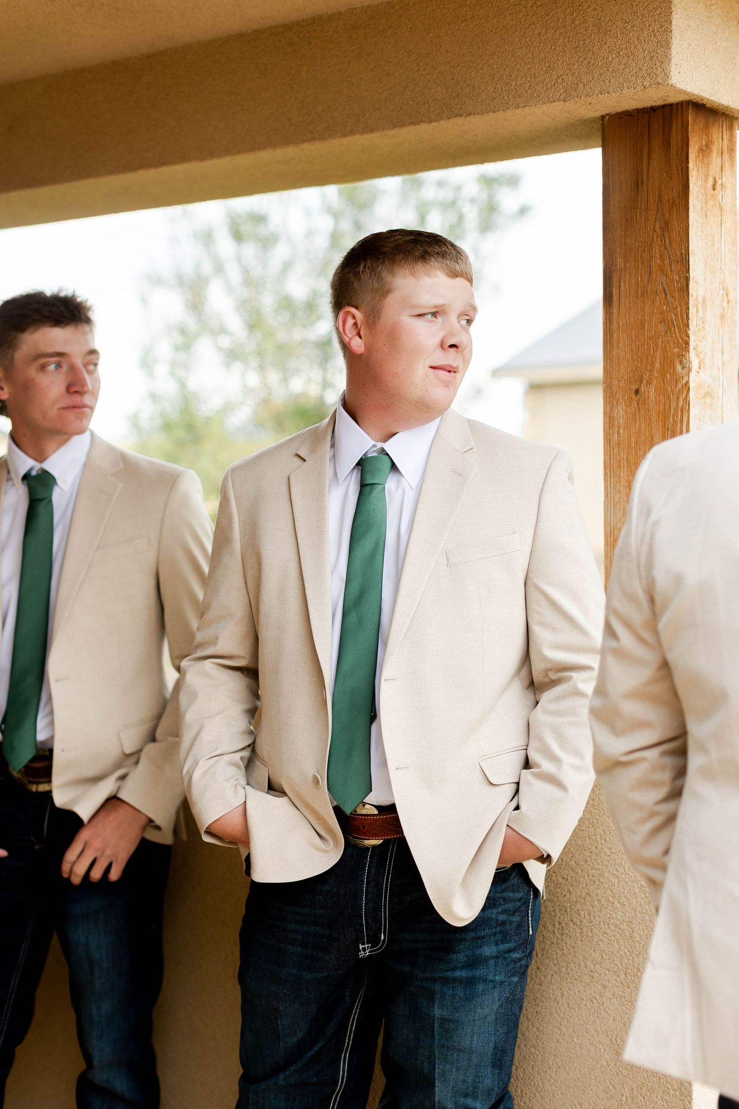 Groomsmen in tan suits with green ties lean against rustic wooden pillars in casual wedding party photo.