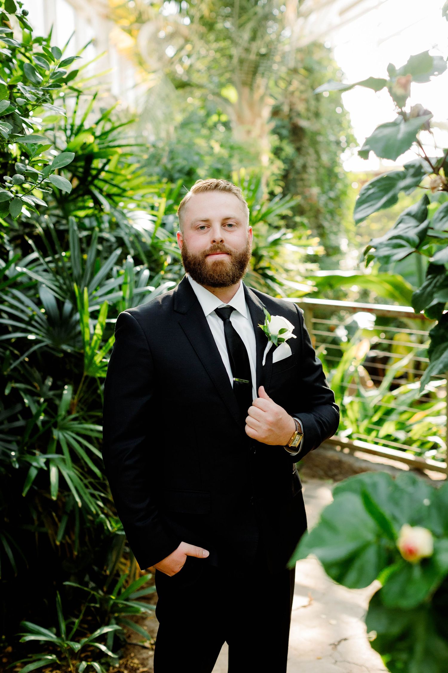 Groom in black suit adjusts his tie while standing among tropical plants.