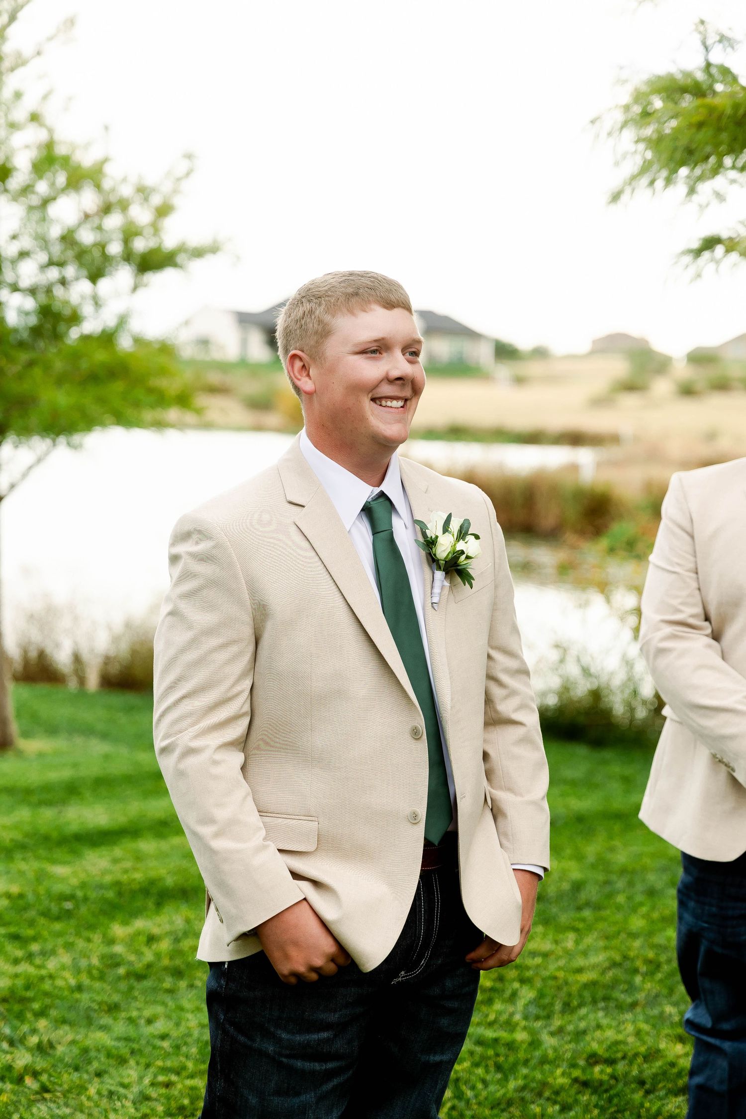 Groomsmen in tan suits and green ties stand together outdoors on a grassy lawn.