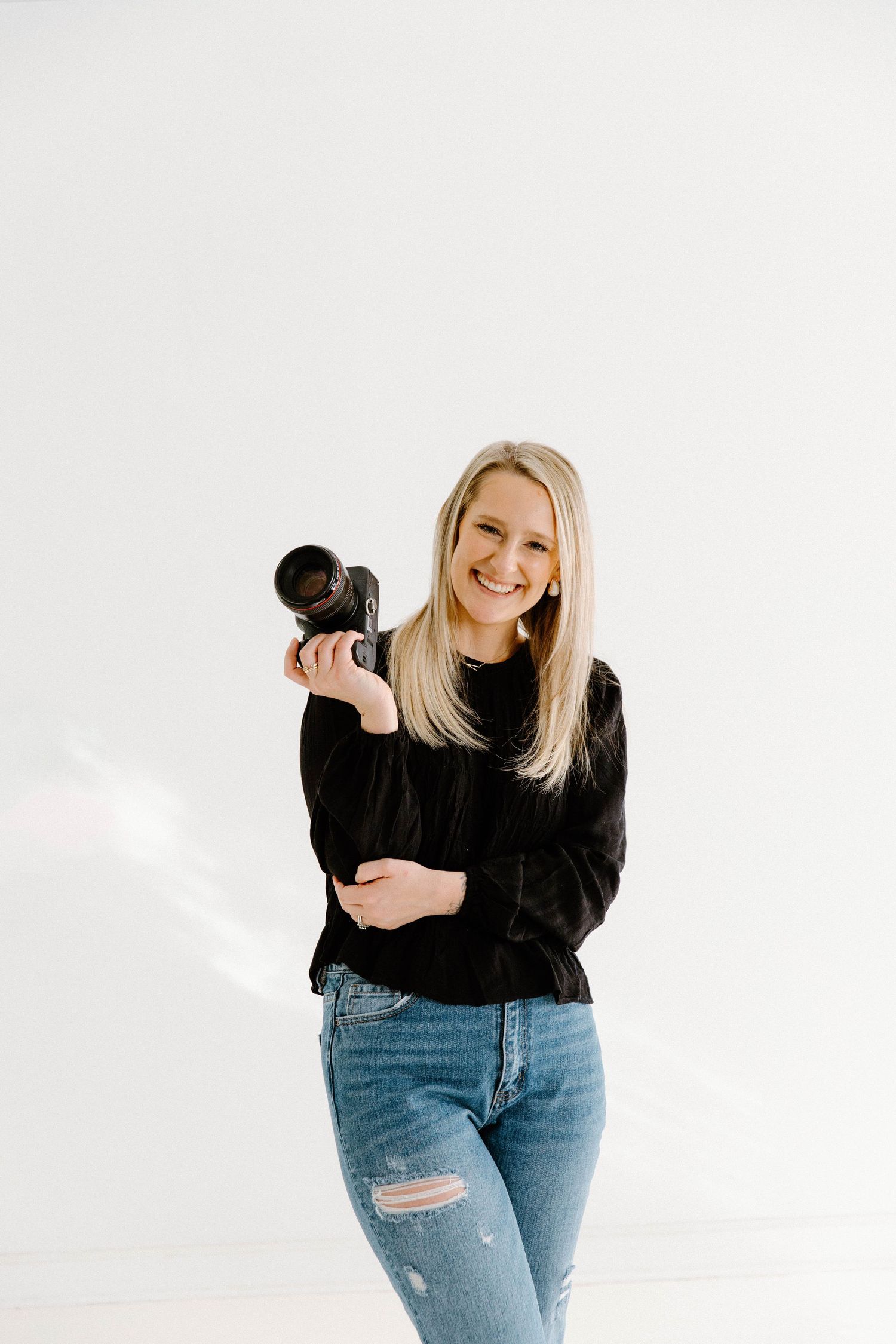 Smiling blonde woman holding a DSLR camera, wearing black top and jeans, posing in a bright white studio.