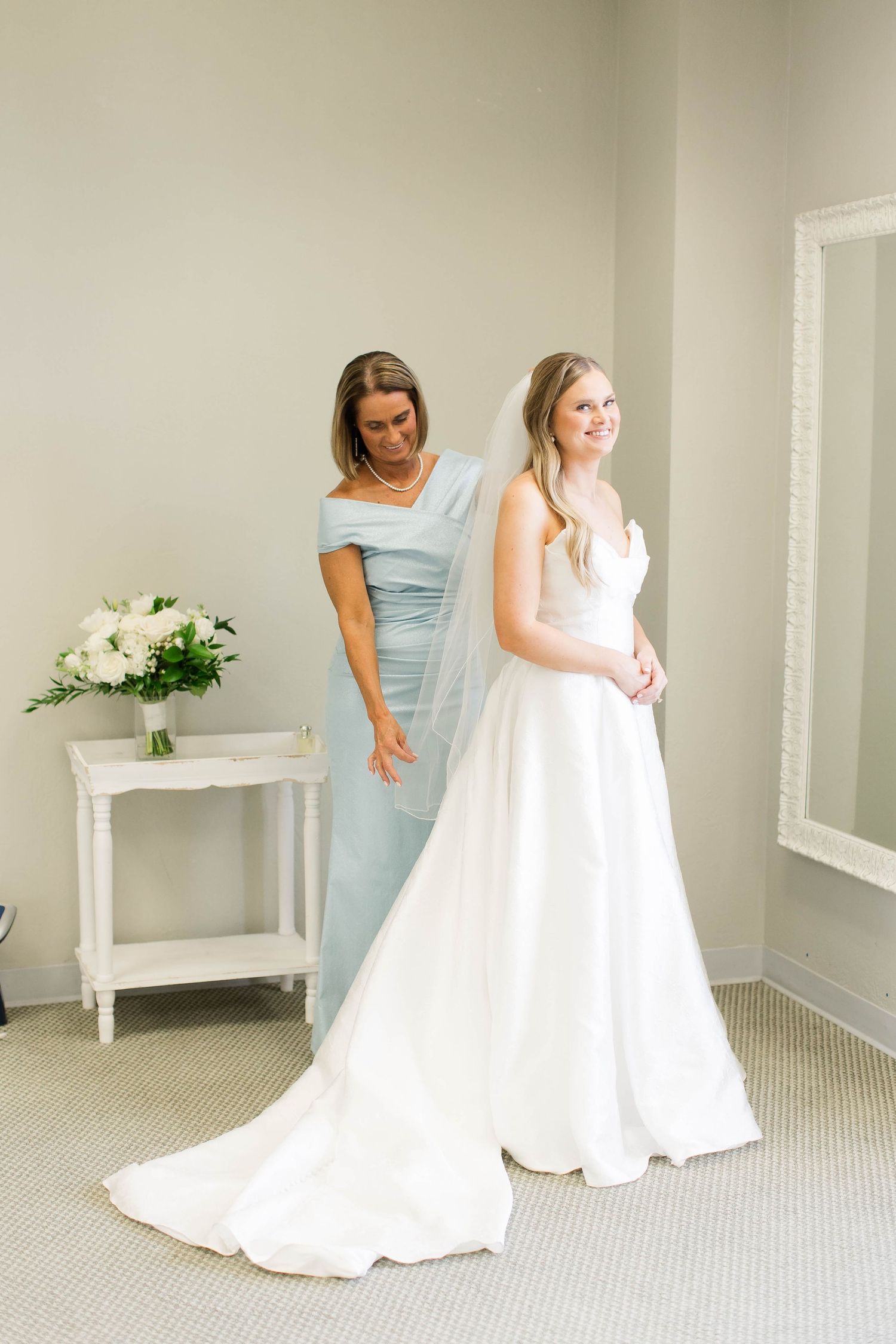 A woman in a white wedding gown gets final adjustments to her dress from an attendant in a light blue dress.