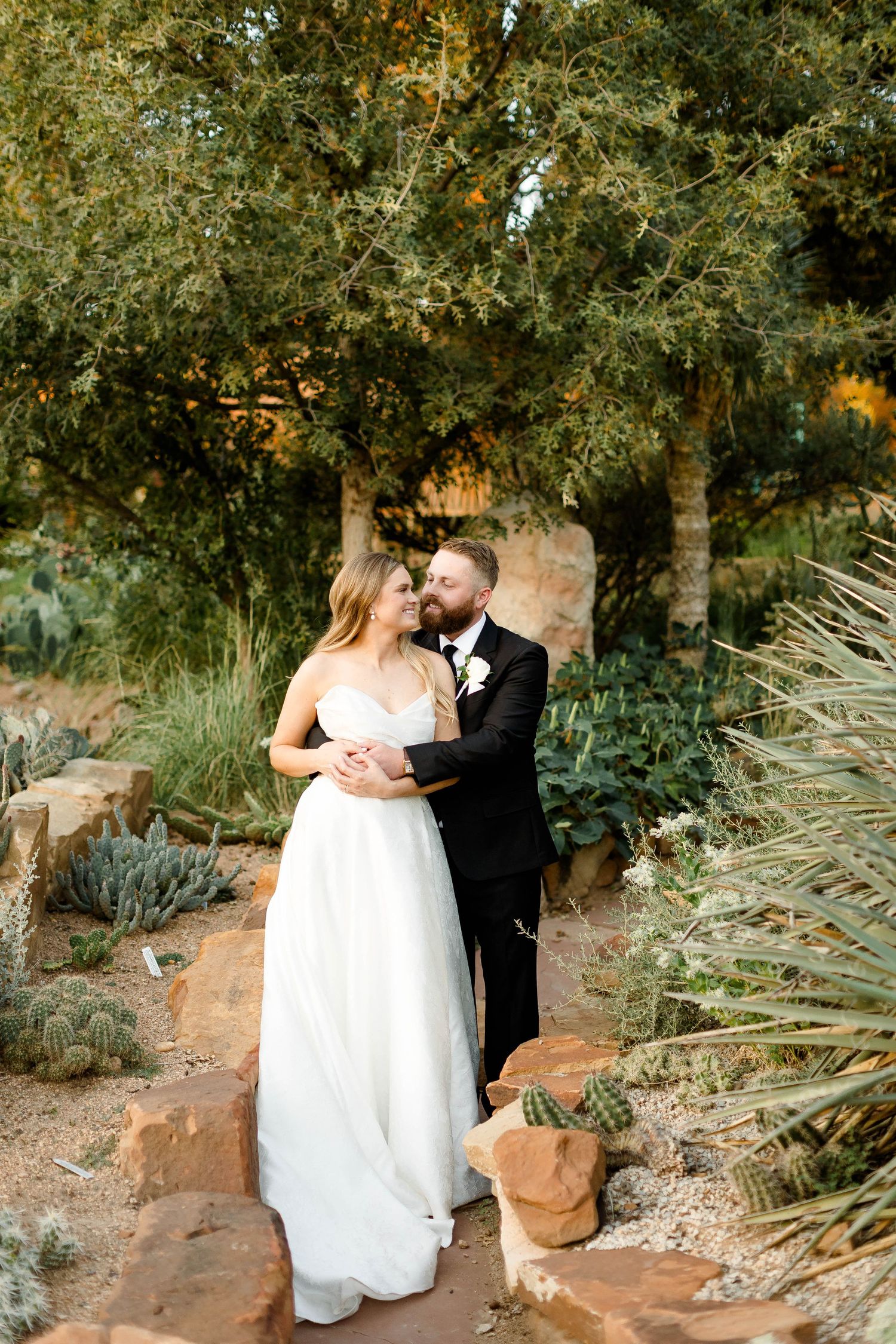 Newlyweds embrace among desert plants and rocks in a garden landscape.