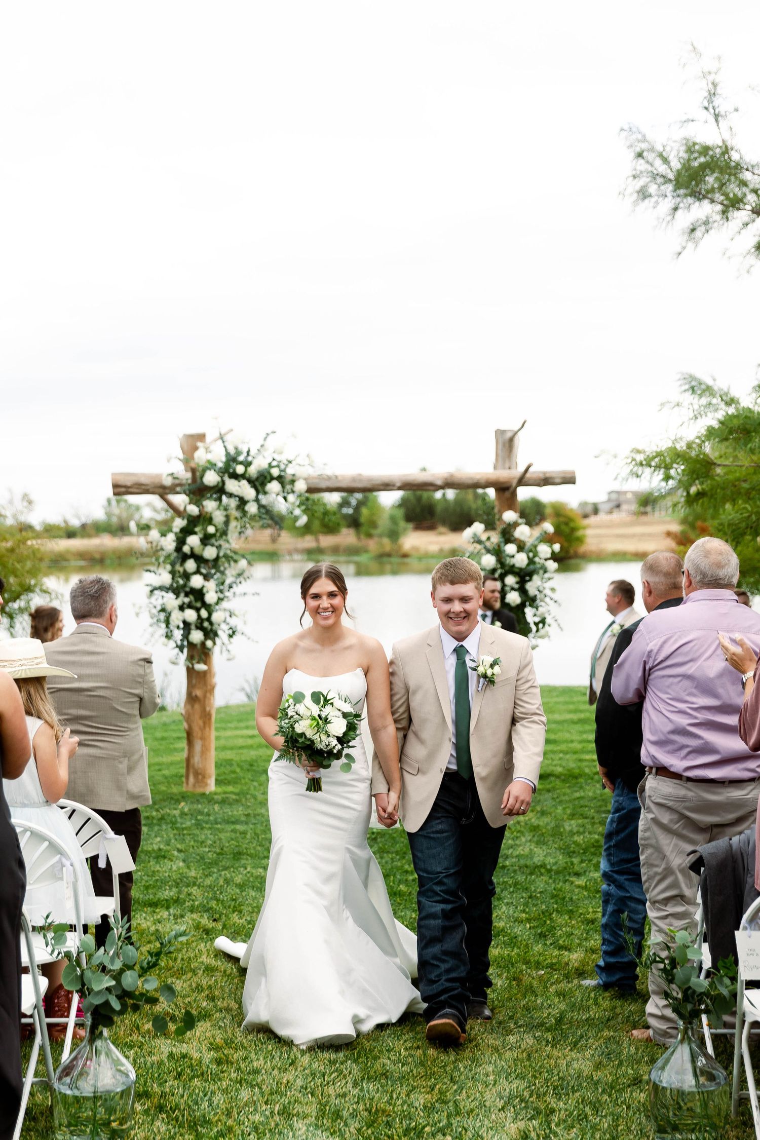 Wedding guests gathered on a lawn watching the ceremony near a rustic wooden arch with white flowers.