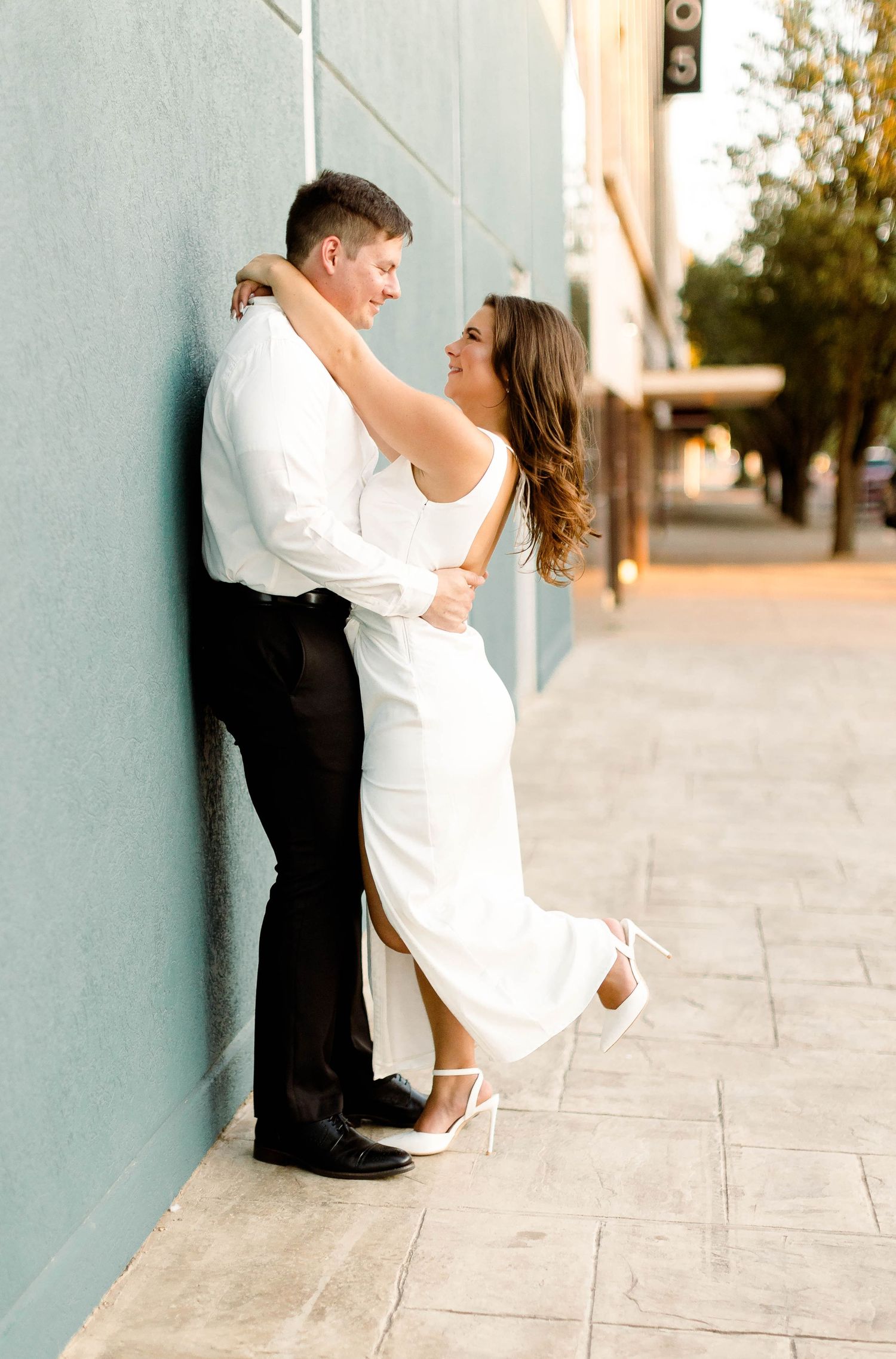 A romantic couple poses against a wall in an outdoor urban setting.