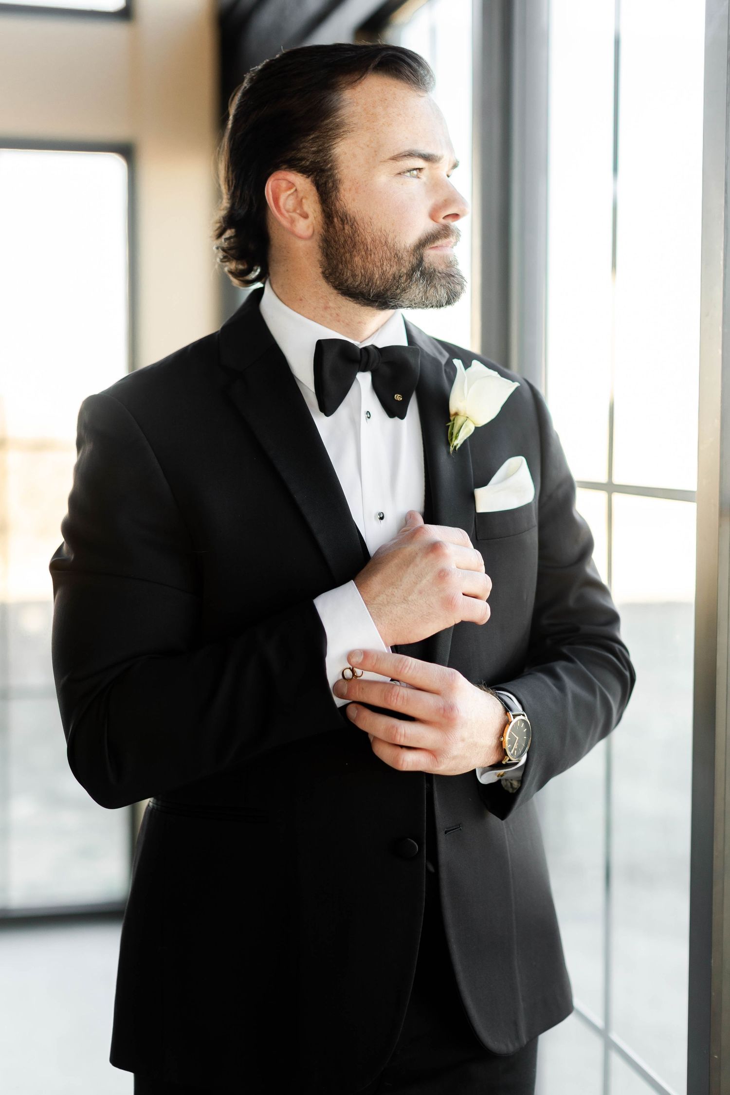 A groom in a classic black tuxedo and bowtie adjusts his cuff by a window.