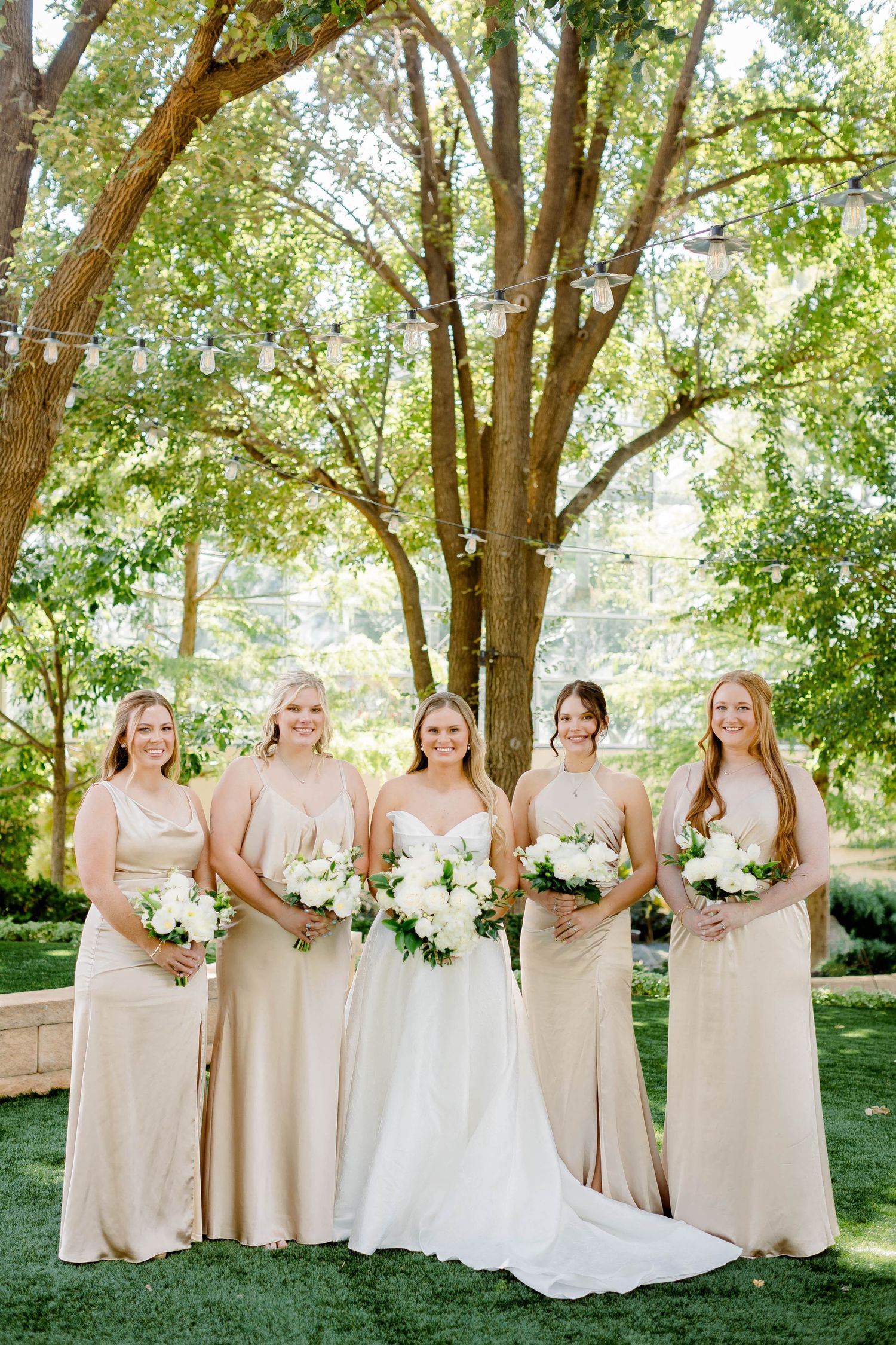 A wedding party in champagne-colored bridesmaid dresses poses together under green trees on a sunny day.