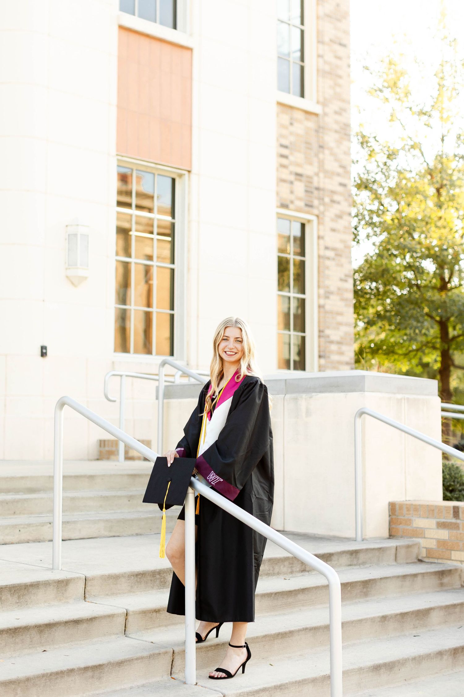 Graduate in black cap and gown poses on concrete steps with white railing.
