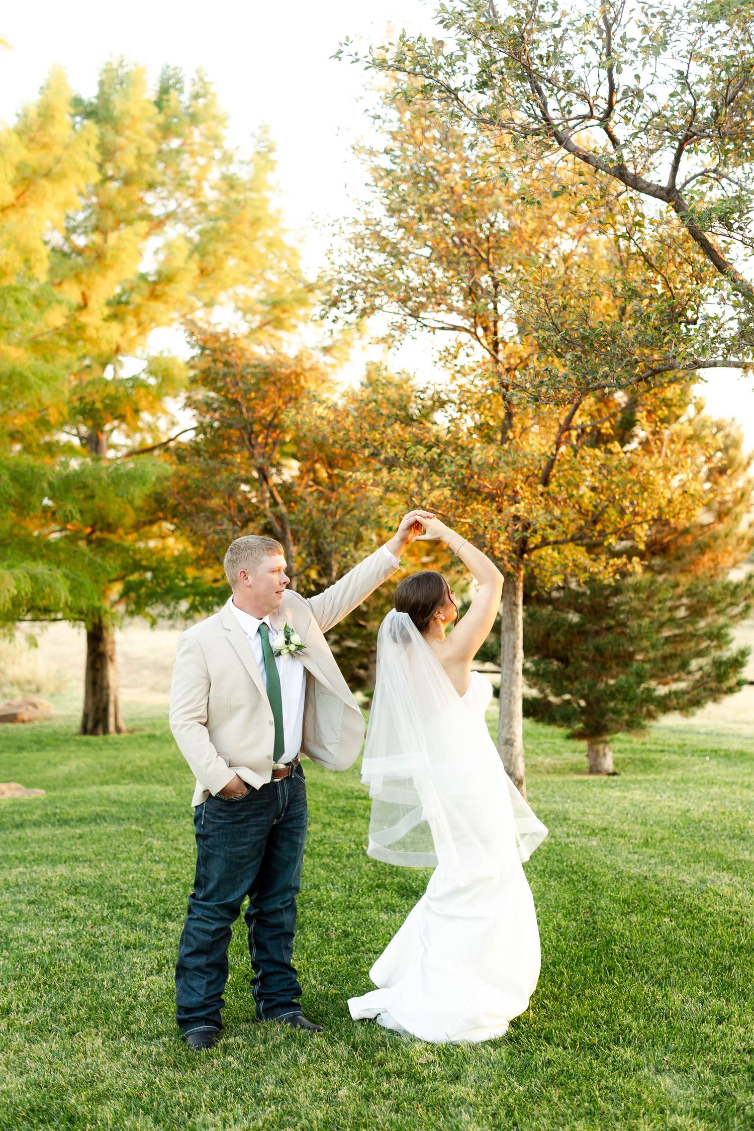 Couple dances playfully on green grass during golden hour with autumn trees in background.
