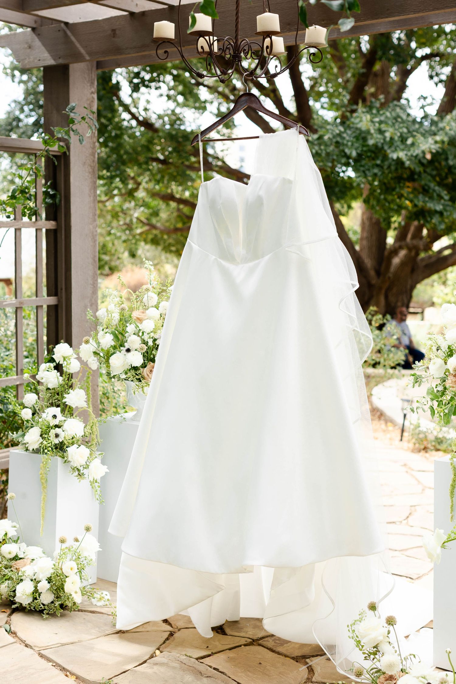 Classic white wedding dress hanging on a garden pergola surrounded by white flowers and greenery.