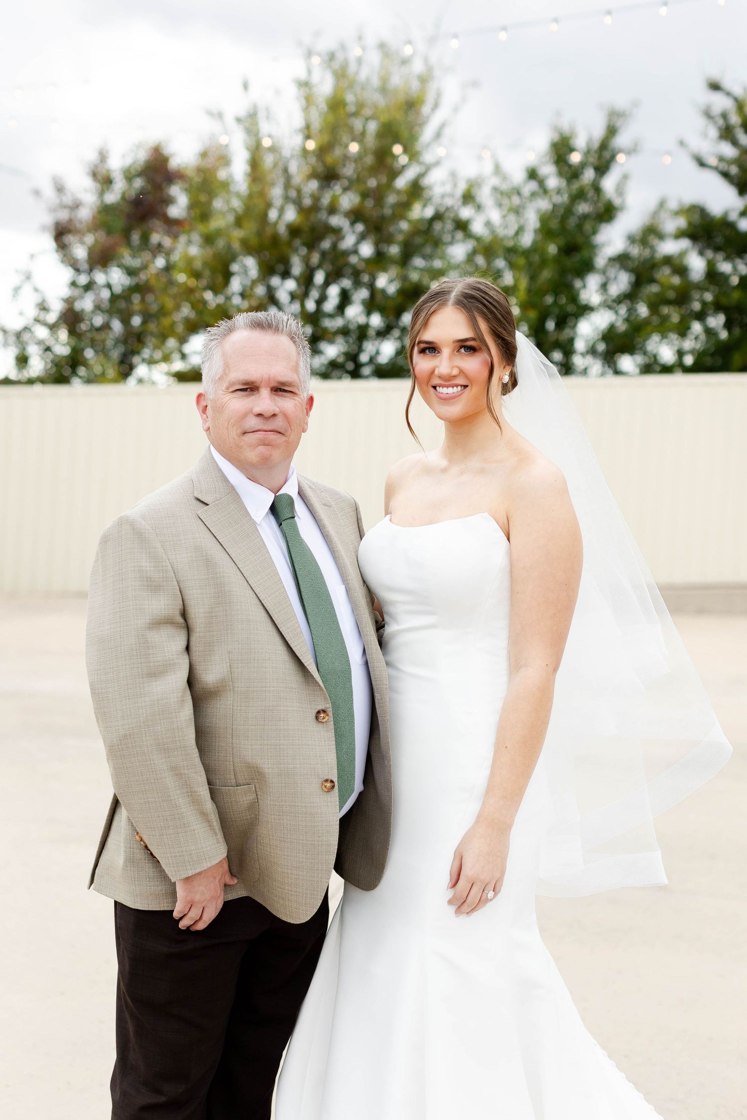 A formal wedding portrait taken outdoors showing two people in elegant attire against a light-colored wall and trees.
