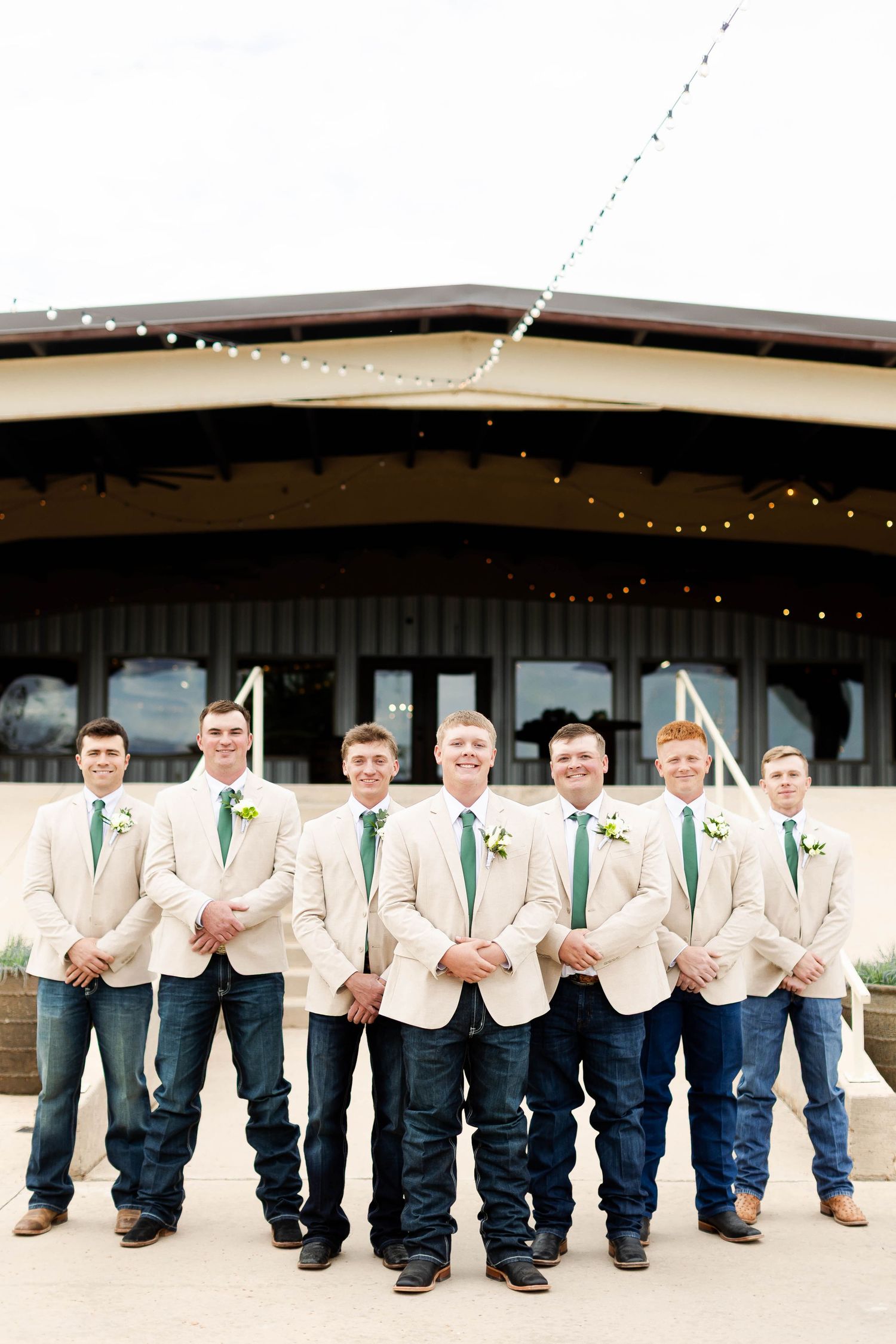 Groomsmen in matching light jackets and jeans pose in a line at a rustic wedding venue with string lights overhead.