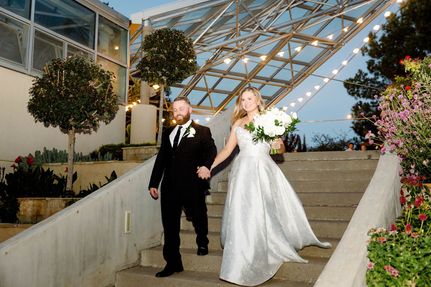 A couple in formal attire walking under a modern glass-roofed architectural structure.