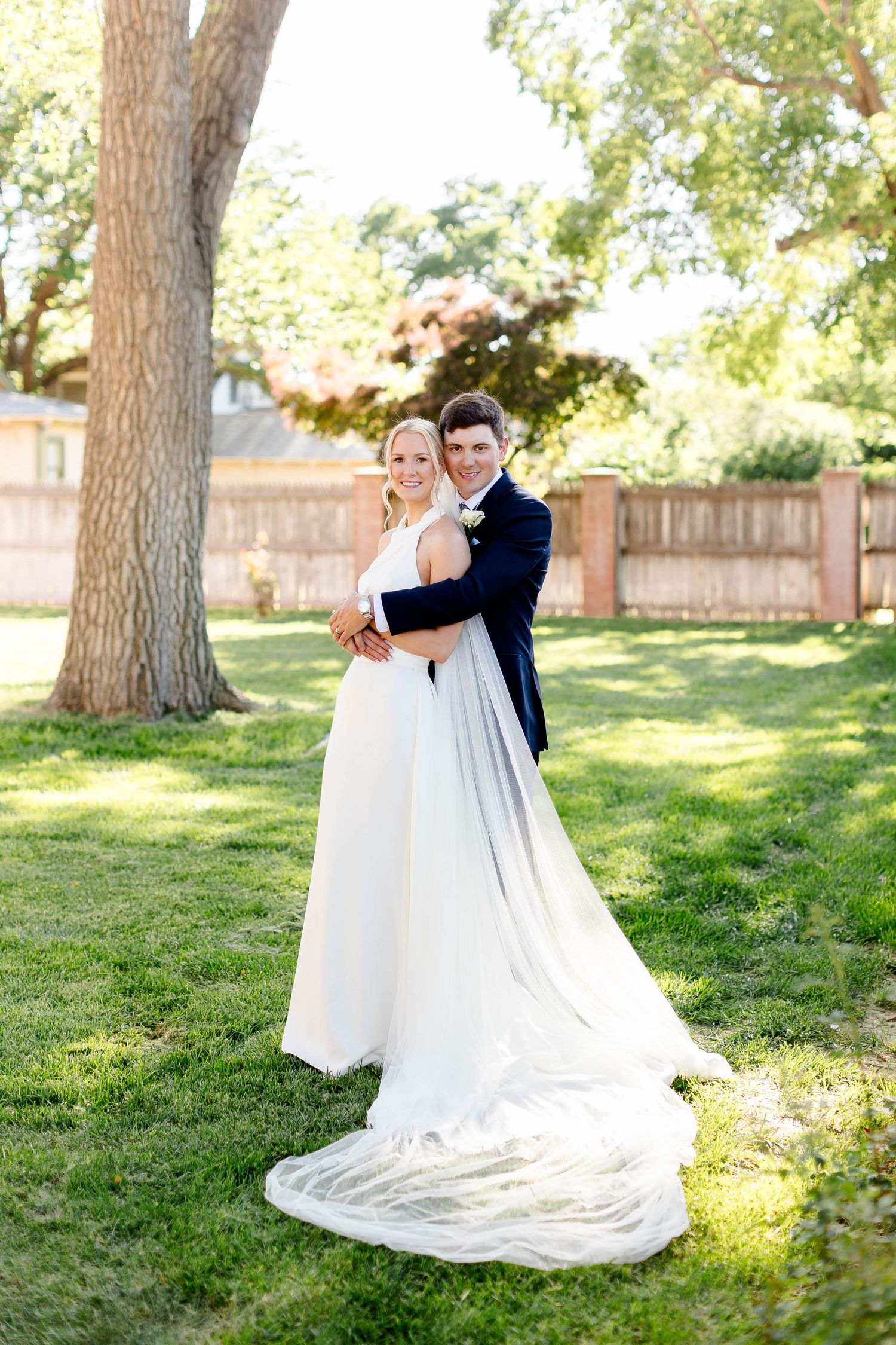 Romantic full-length portrait of newlyweds embracing in a sunny garden with flowing wedding dress train.