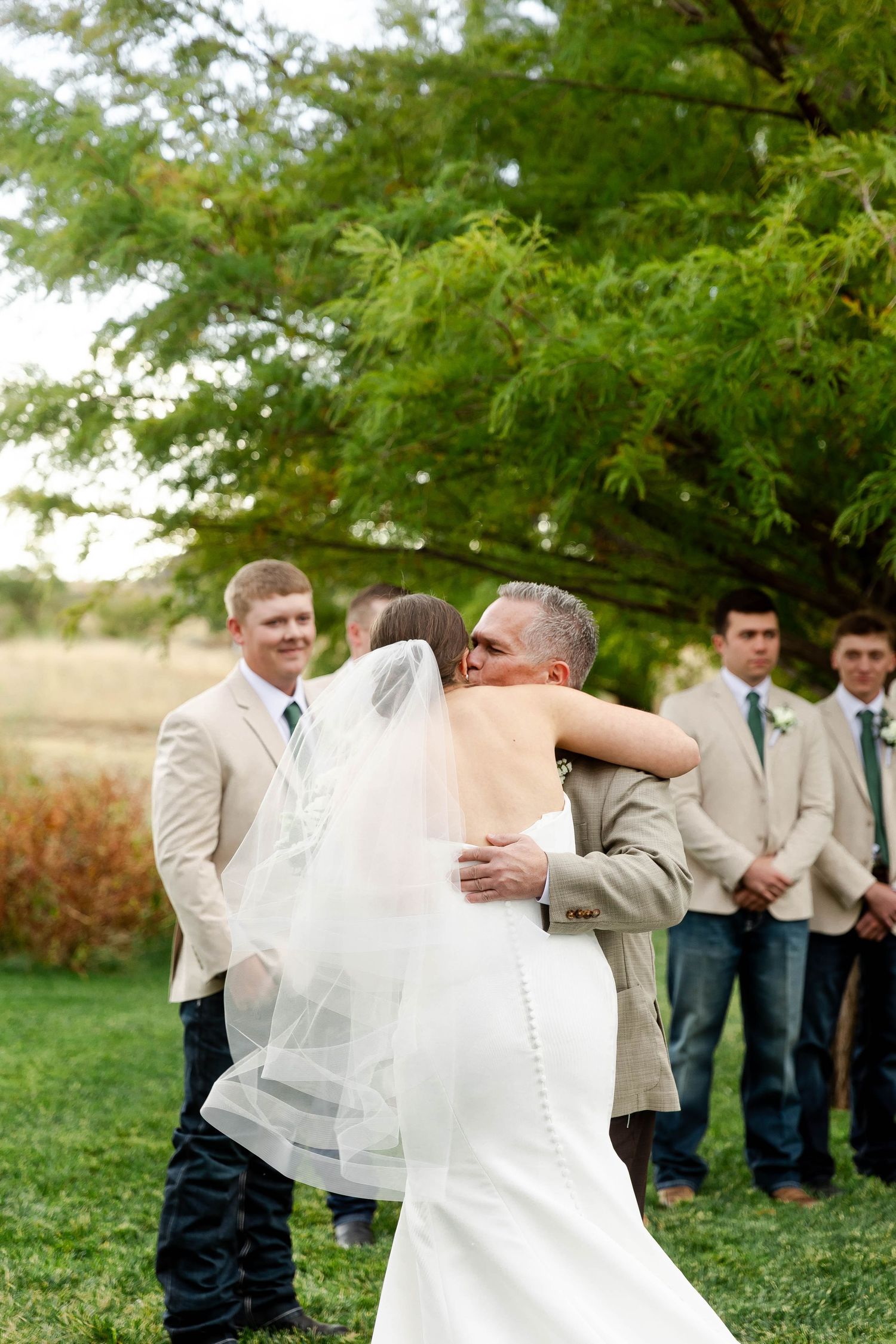 Wedding party in light colored attire gathers on a grassy lawn under green trees.
