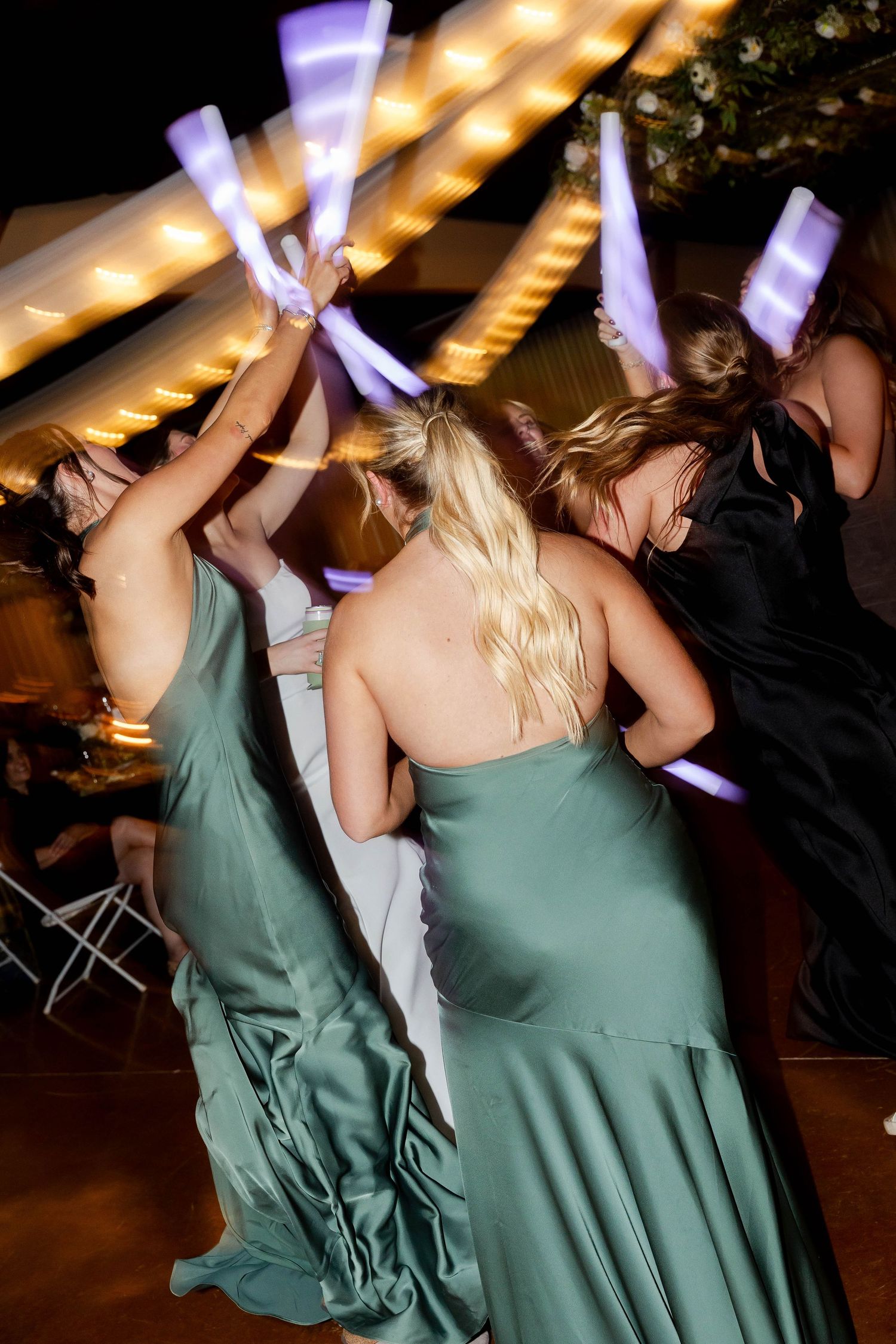 Bridesmaids in sage green dresses dance with raised arms at a dimly lit wedding reception.