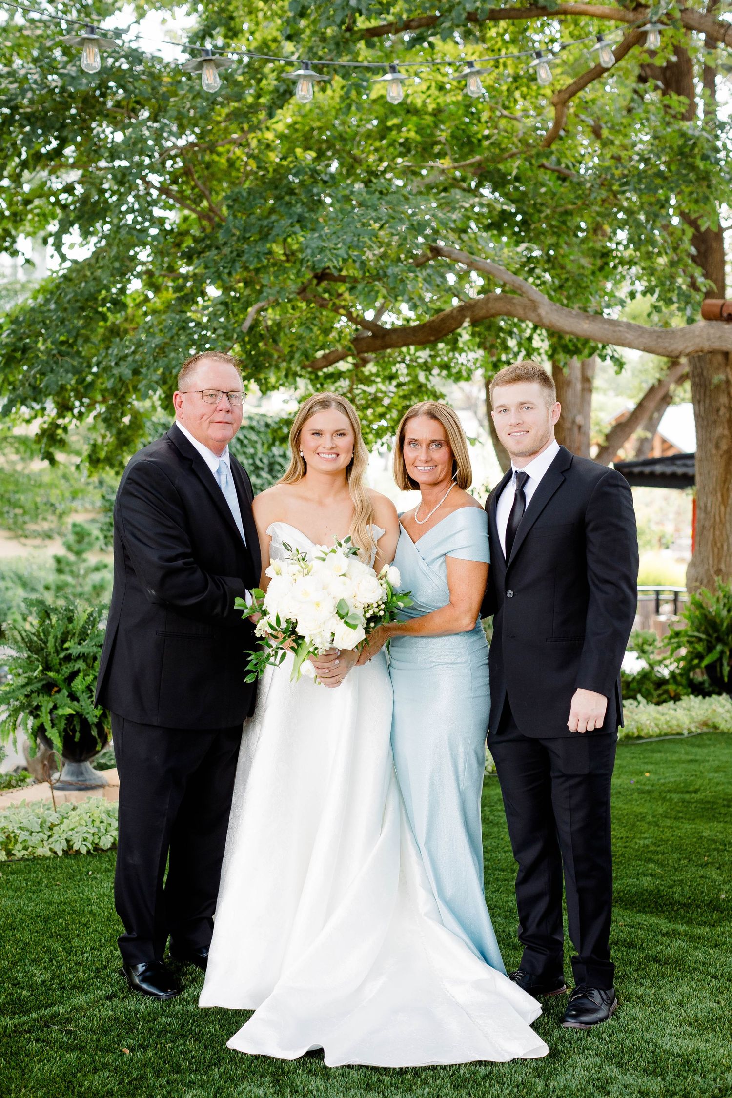 Wedding party poses together on manicured lawn with trees in background.