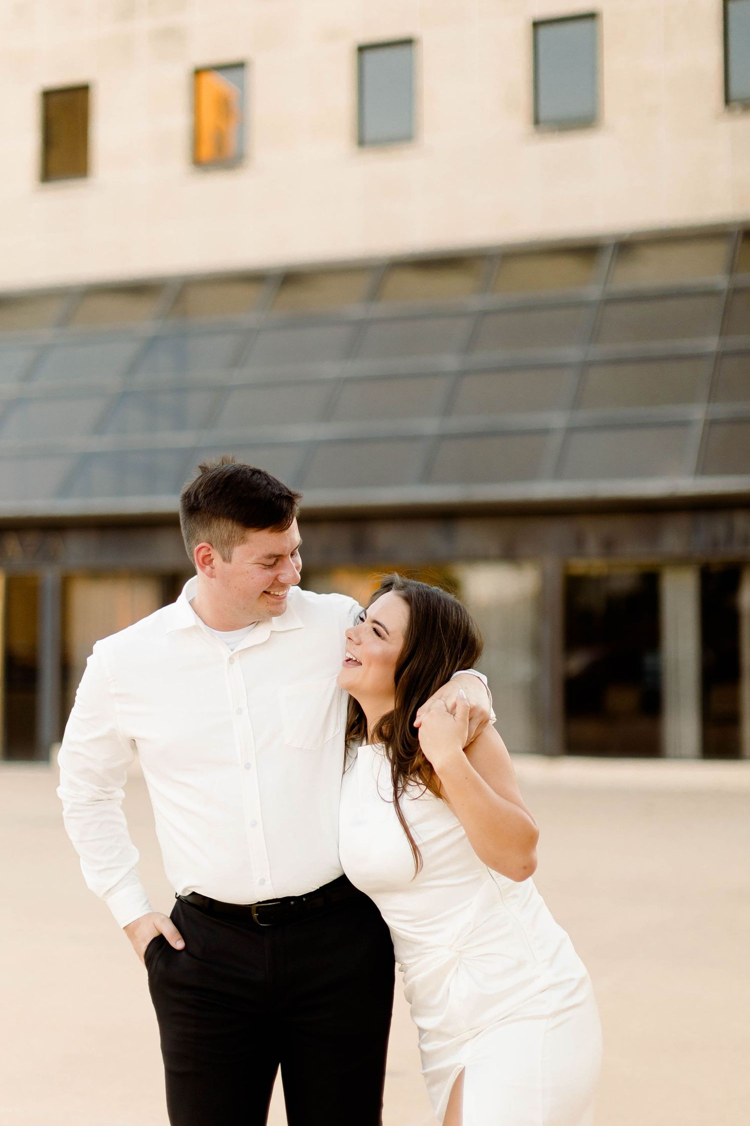 A couple in formal wear shares a playful moment outside a modern building.