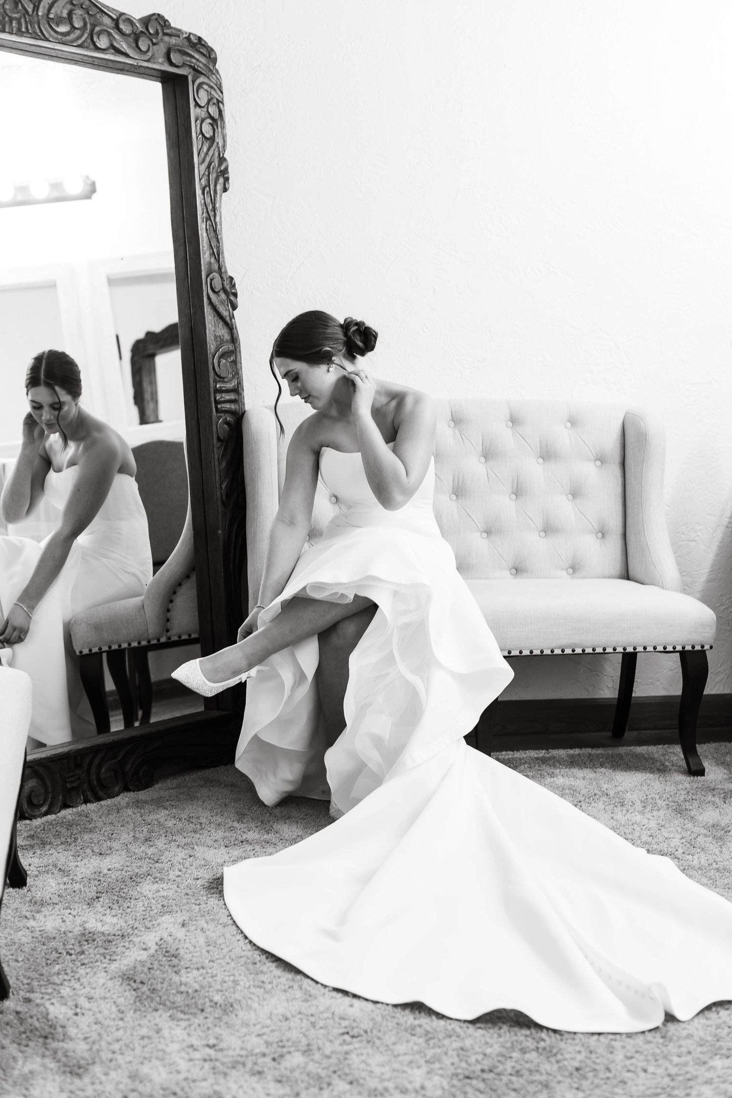Black and white photo of a bride in a flowing white dress putting on her shoes while sitting on an upholstered bench.