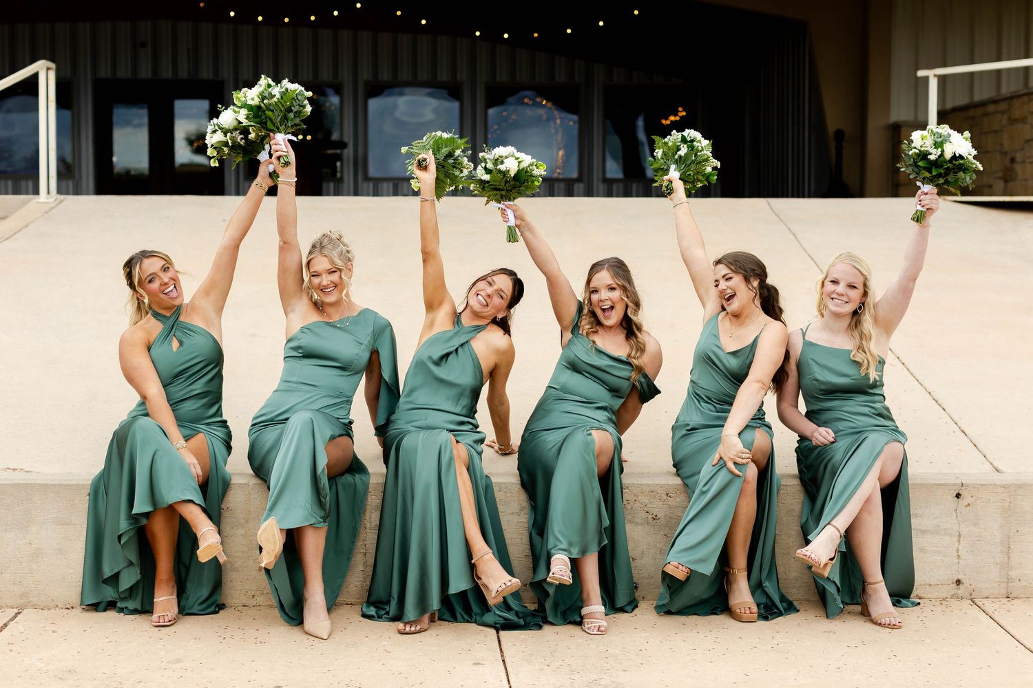 Row of bridesmaids in matching teal dresses sit on stone steps and pose playfully for photo.