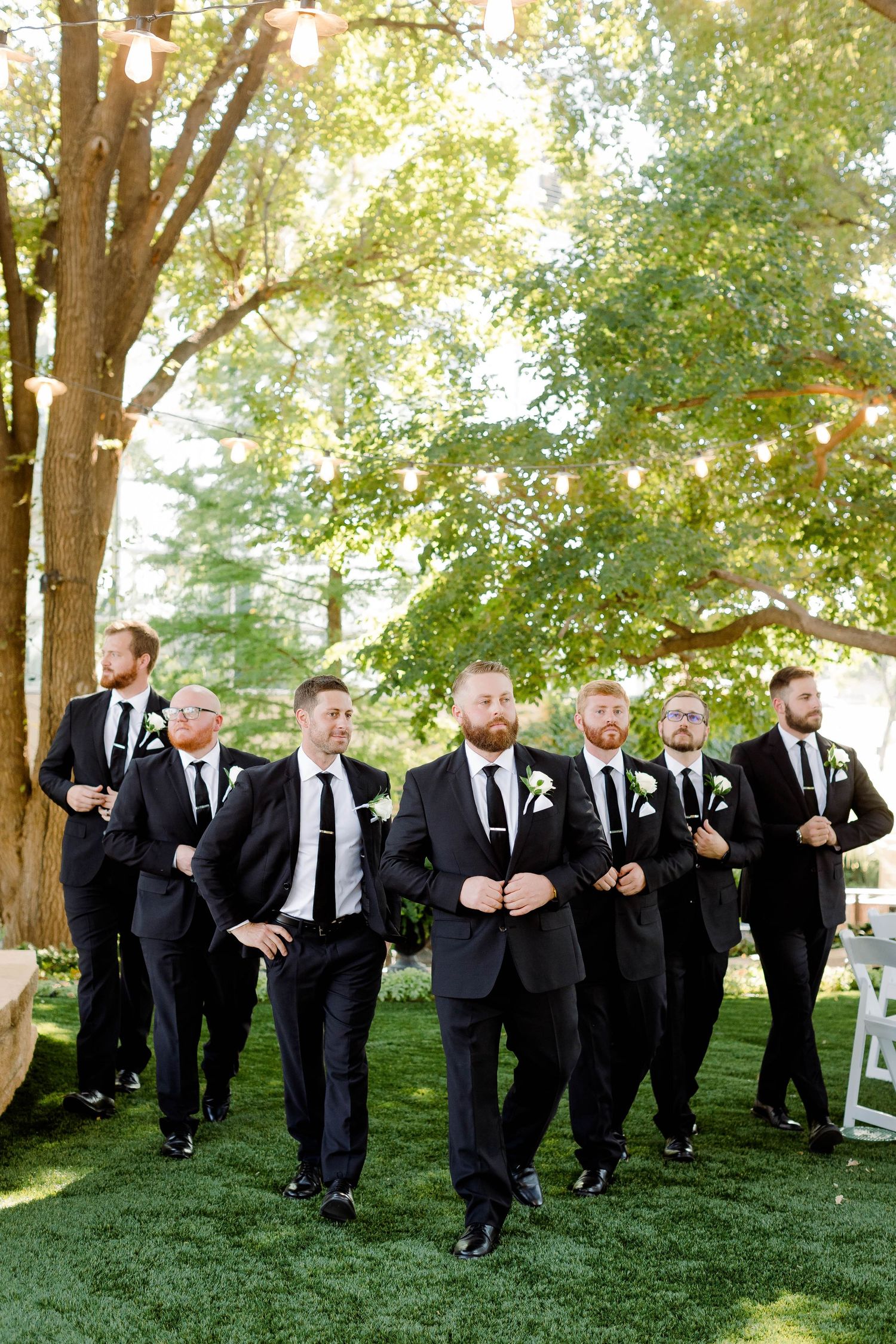Large wedding party in black suits poses energetically on a grassy hill under trees.