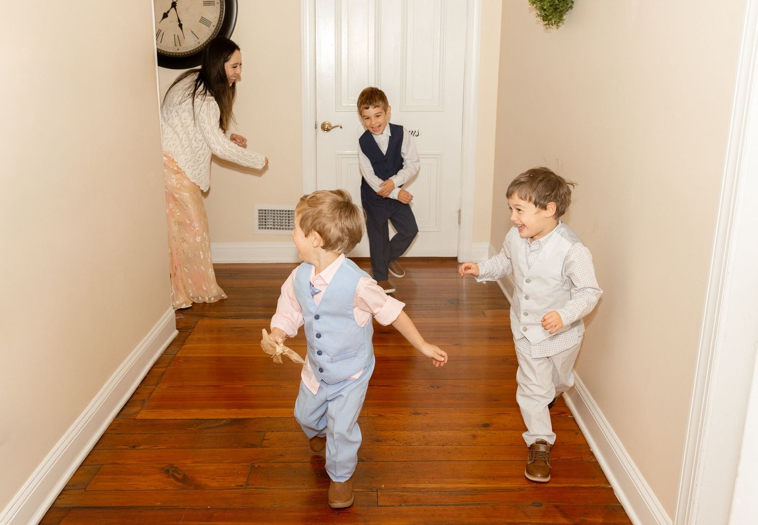 Children playing and running in a hallway with hardwood floors while a woman watches and smiles nearby.