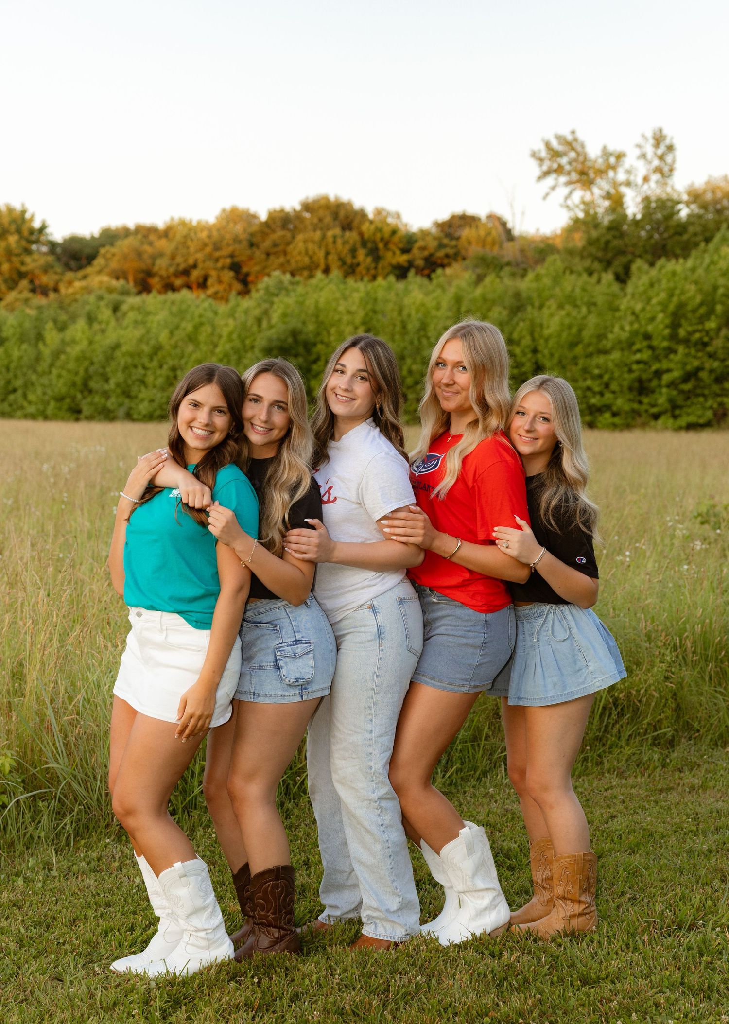 Five young women pose together smiling in a green field, wearing casual summer outfits including boots and denim shorts.