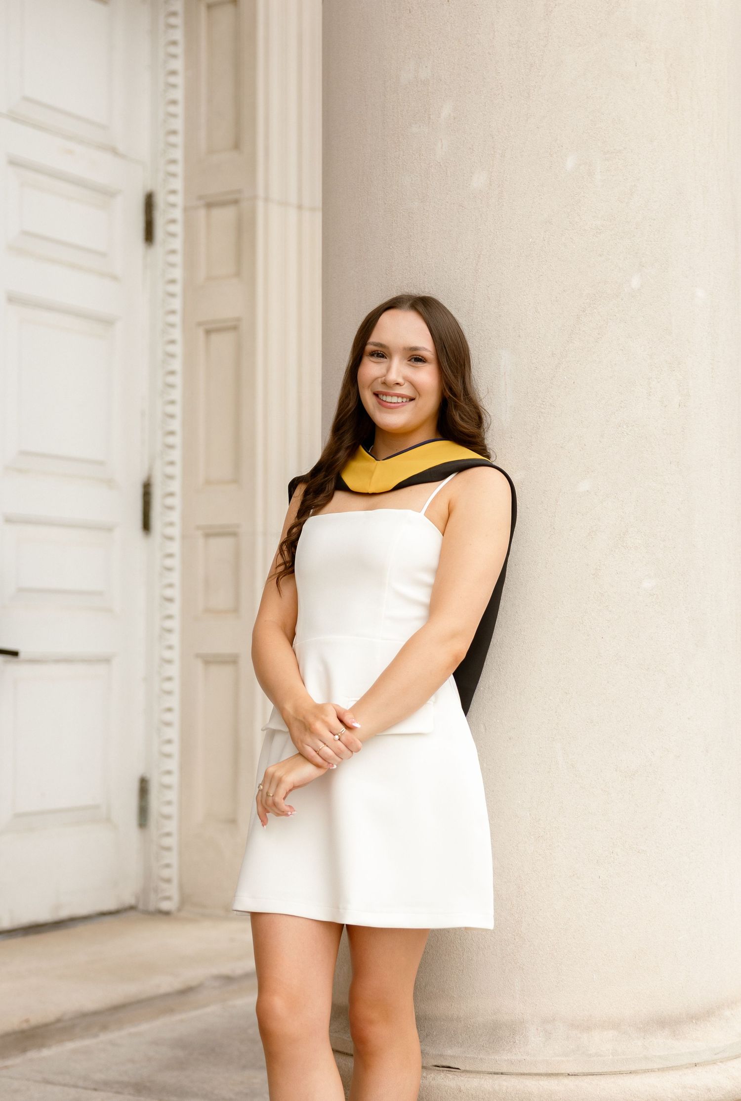 Smiling young woman in white dress with gold graduation stole leaning against a white column outdoors.