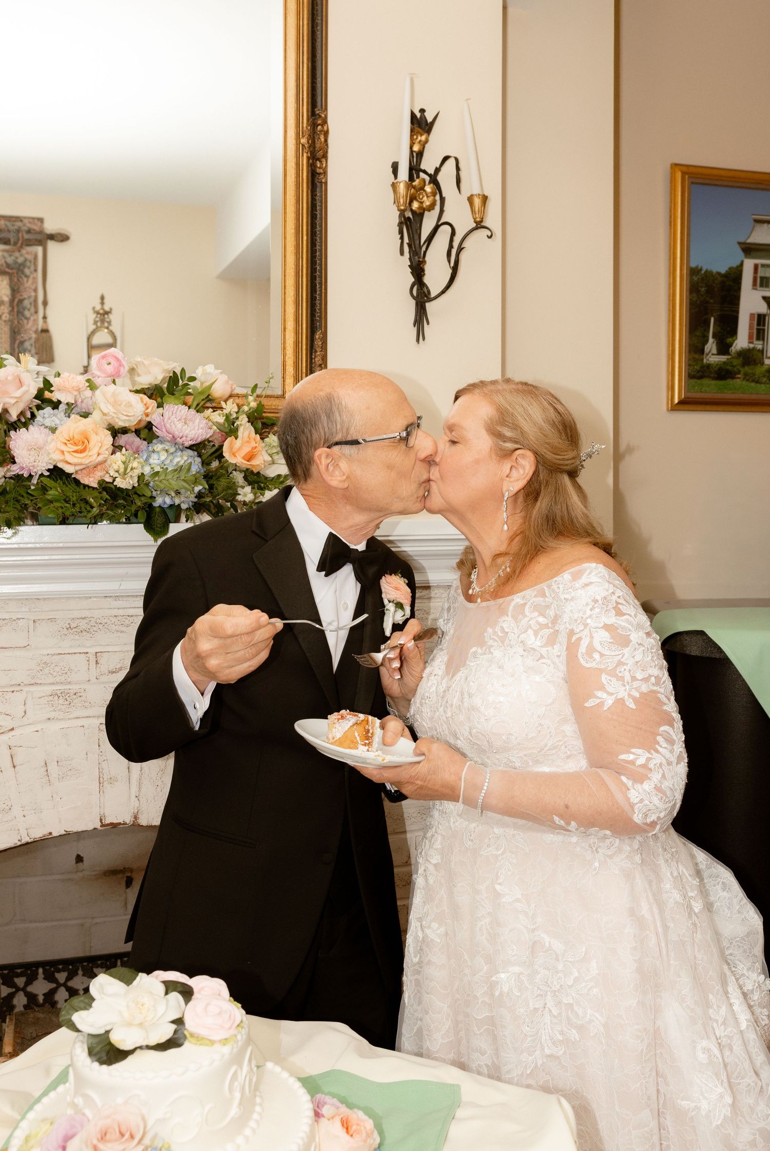 Elderly couple sharing a kiss while cutting wedding cake, bride in lace gown, groom in tuxedo, floral arrangement backdrop.