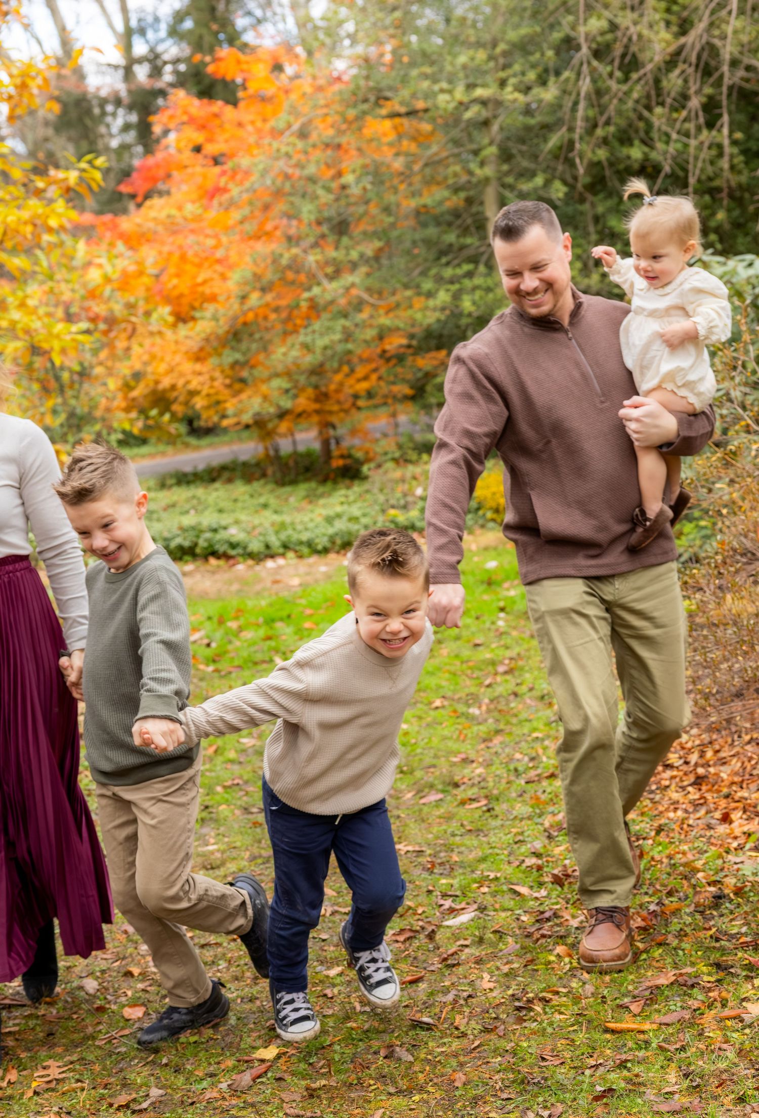 Family walking outdoors in autumn park, dad carrying baby girl while two boys play, colorful fall foliage in background.