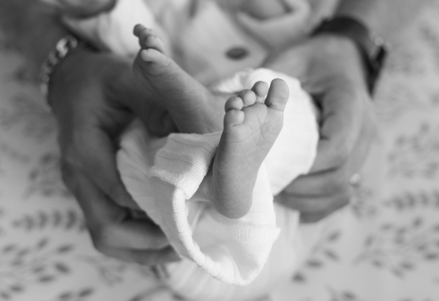 Black and white close-up of tiny baby feet being gently held by adult hands against a textured background.
