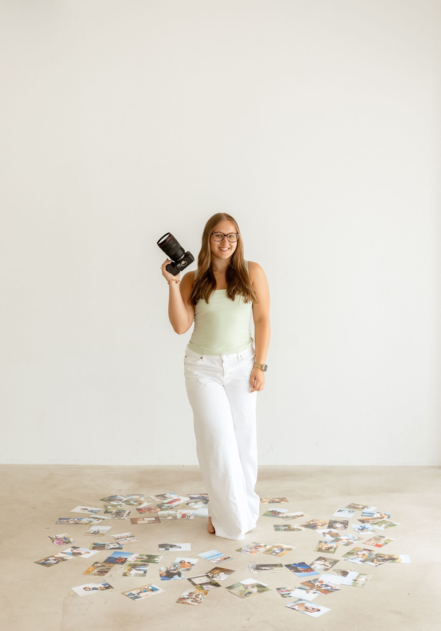 Woman holding camera surrounded by scattered photos on floor in bright minimalist studio, wearing white pants and green top.