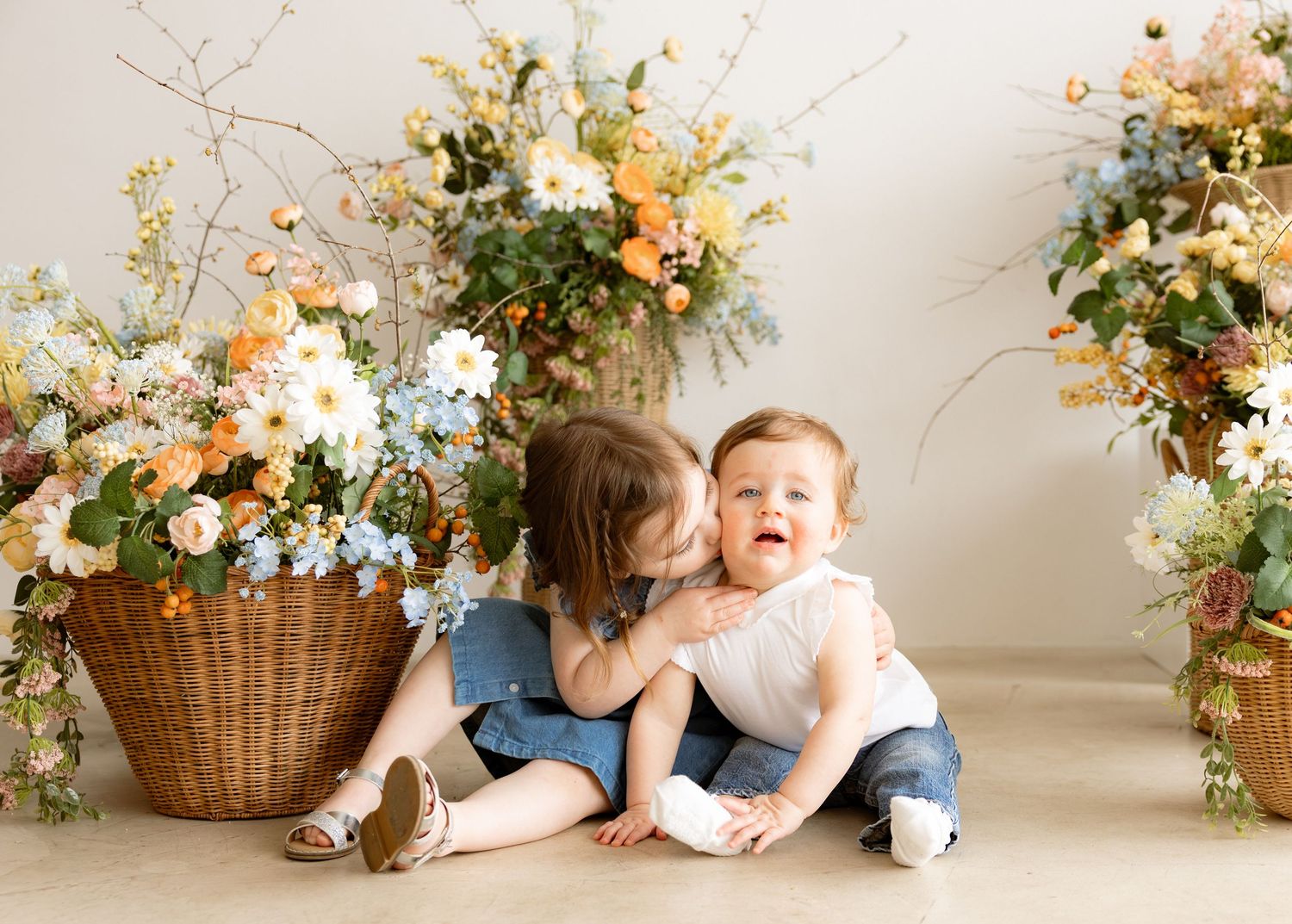 Older toddler girl kissing baby brother on cheek surrounded by floral arrangements in a bright studio setting.
