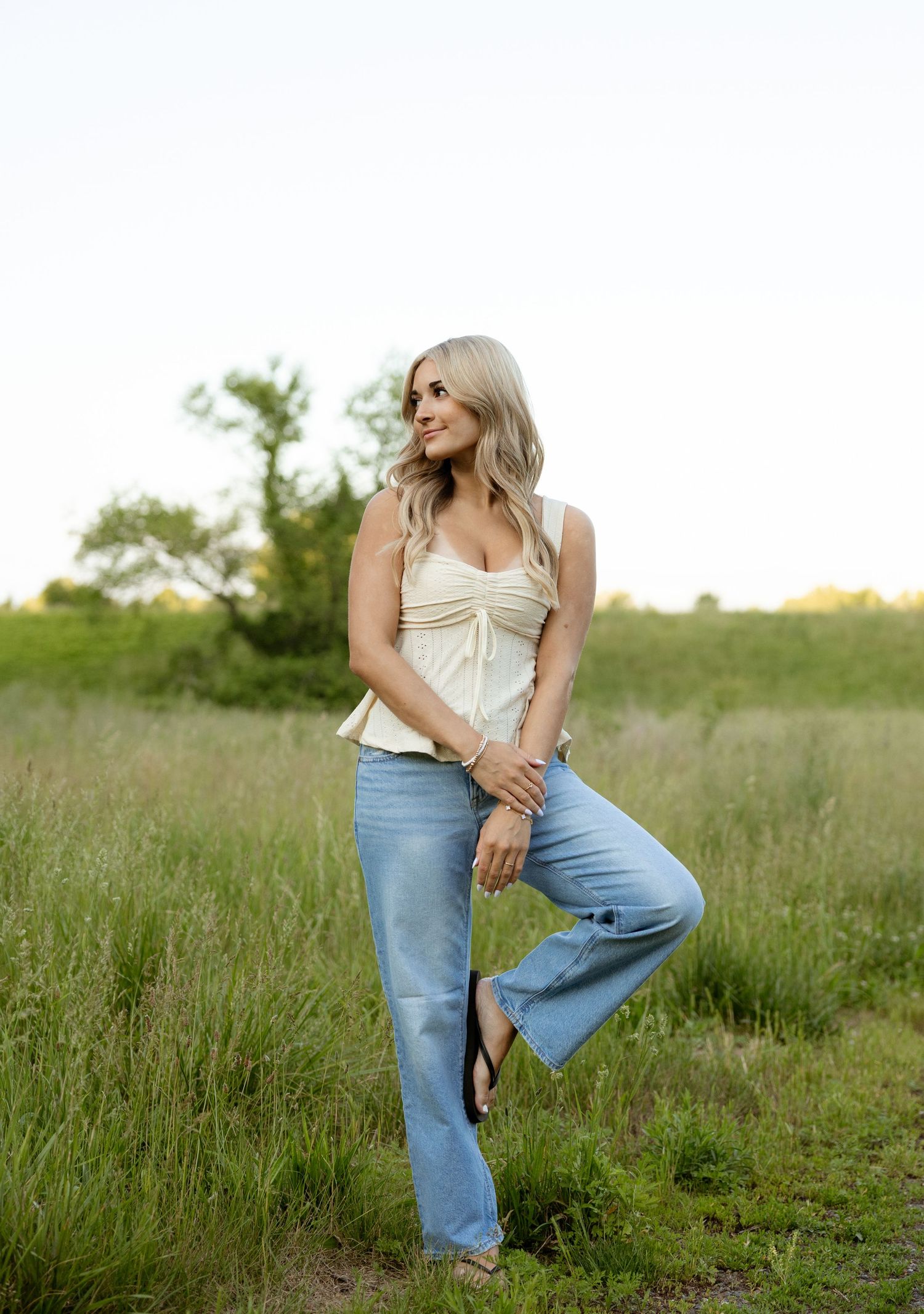 Blonde woman in cream top and flare jeans posing in a green meadow field during golden hour.