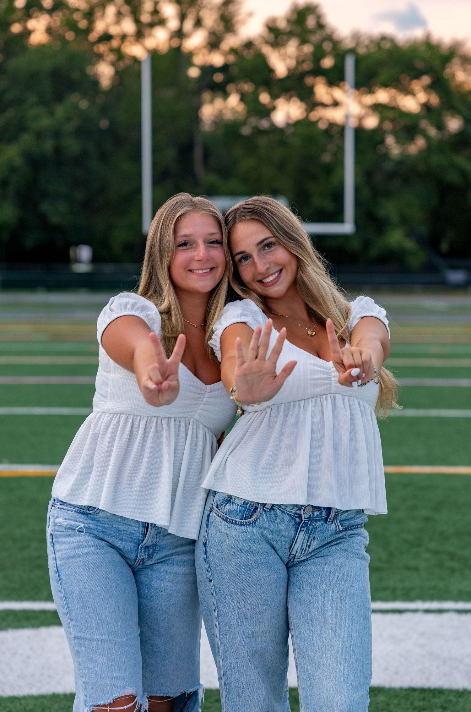 Two friends in matching white tops and jeans pose playfully outdoors at sunset.