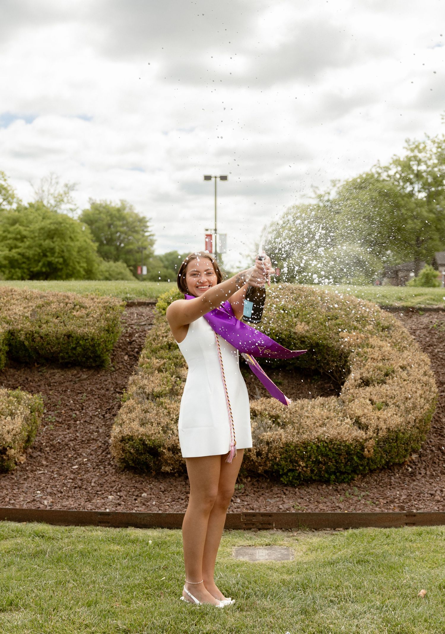 Young woman in white dress and purple sash celebrates outdoors, spraying champagne near decorative hedges.