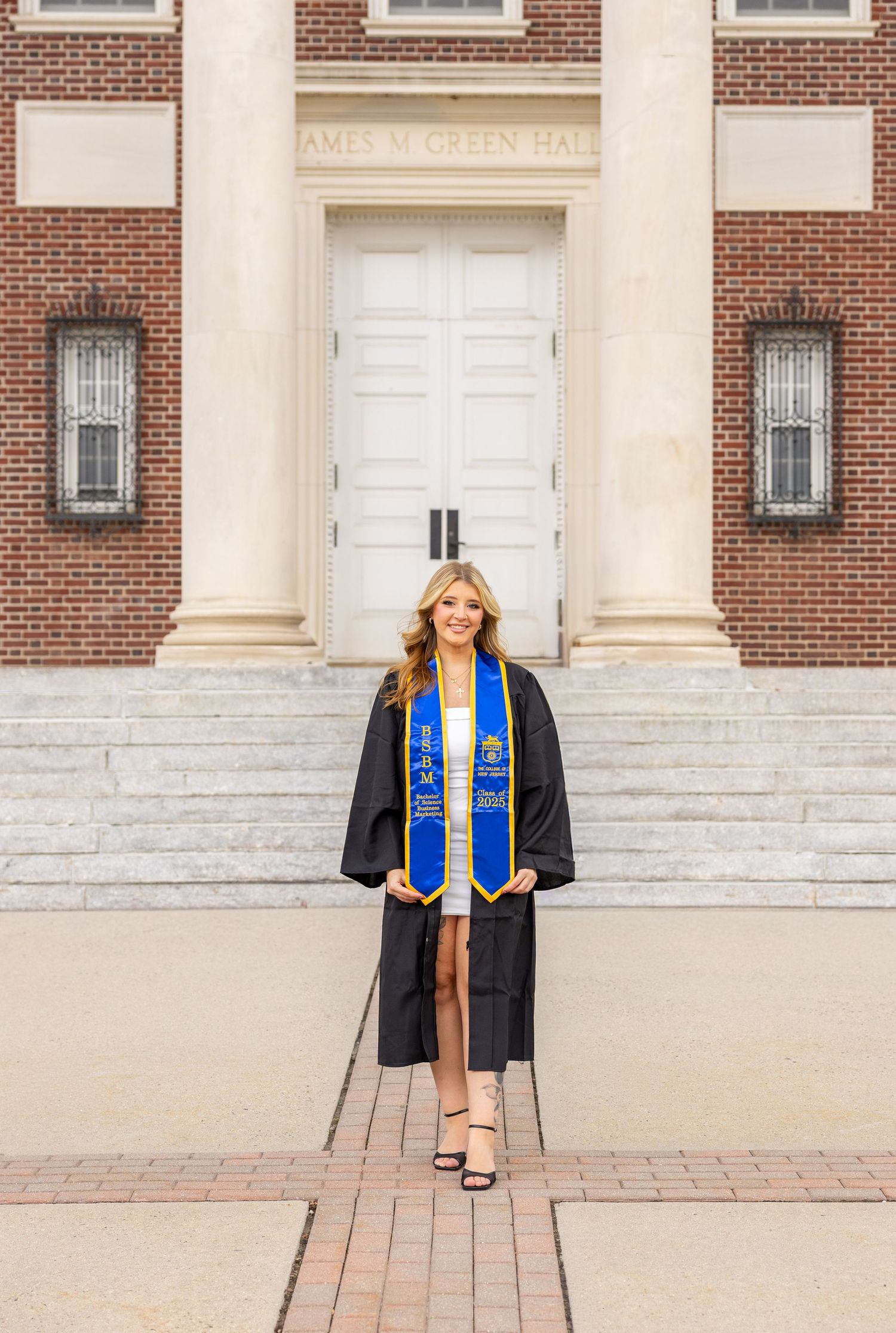 Graduate in cap and gown poses in front of white columned entrance to McLin Hall.
