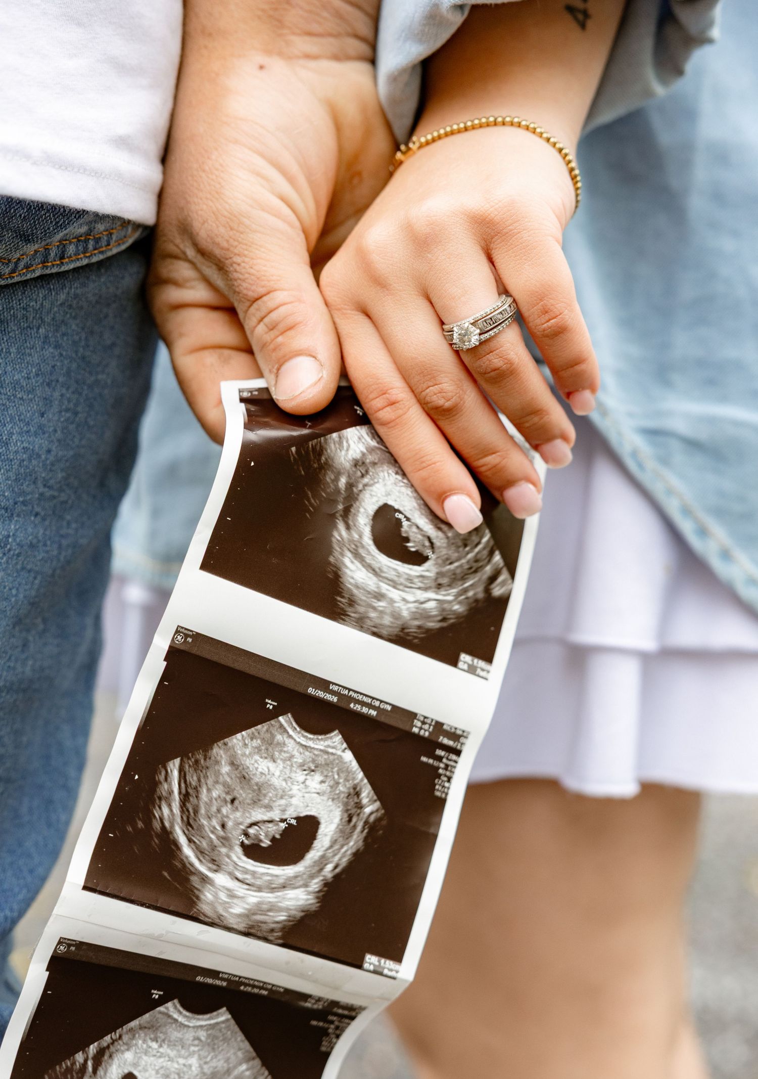 Couple holding ultrasound sonogram photos together, woman wearing engagement ring and gold bracelet, pregnancy announcement.