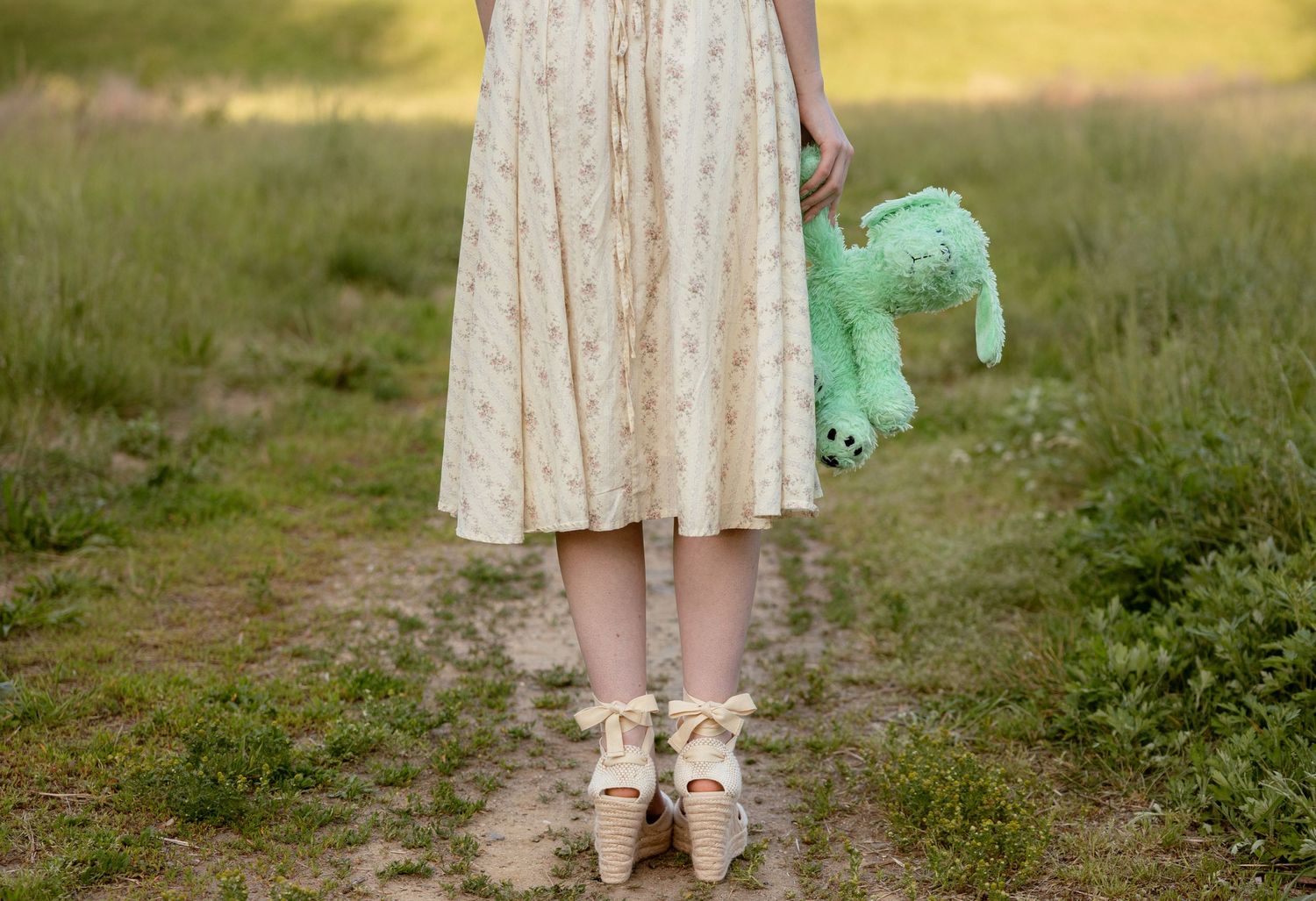 Girl in floral dress and wedge shoes holding green stuffed animal toy on a dirt path in a field.
