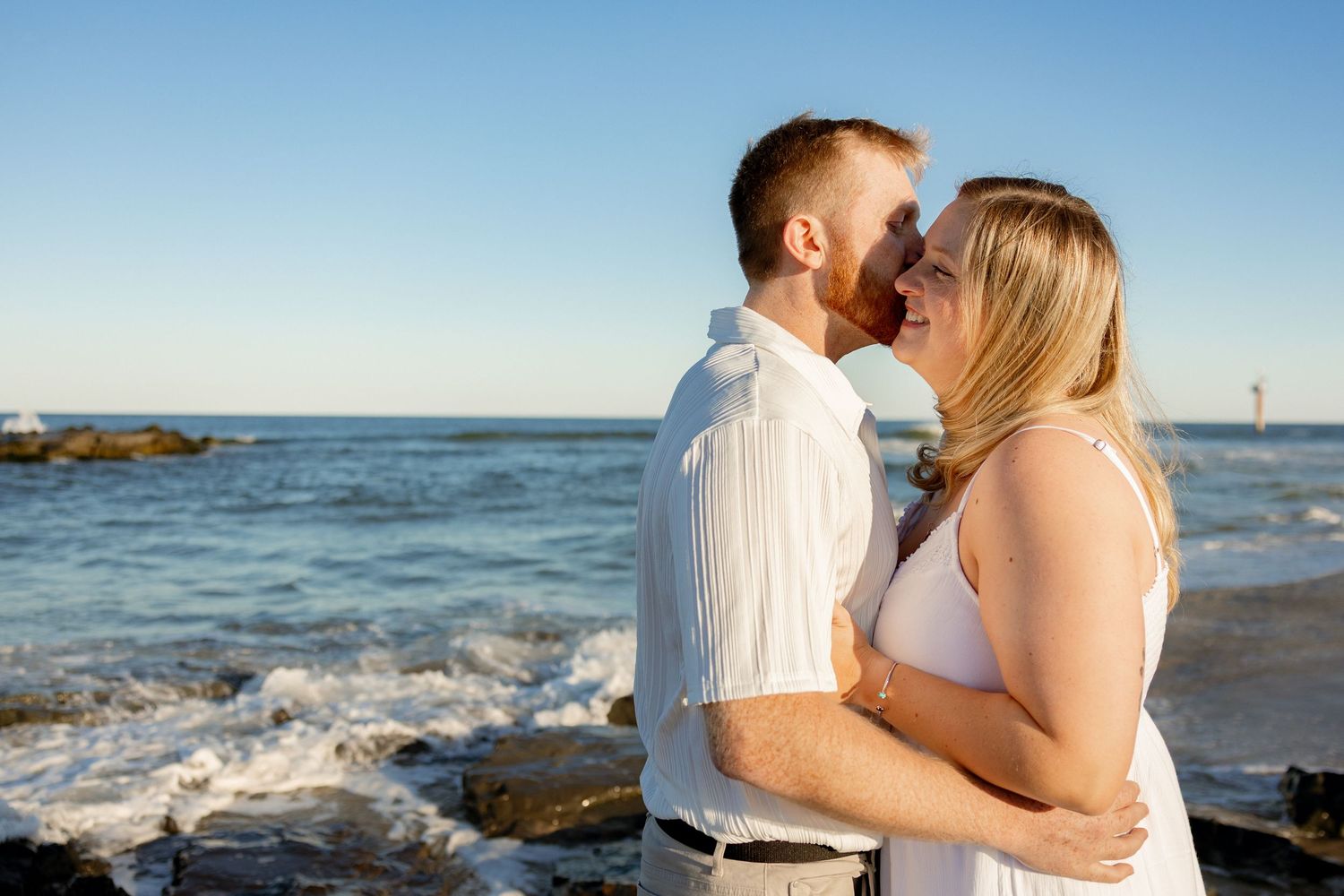 A couple shares a romantic kiss on a rocky beach with blue ocean waves and clear sky behind them.