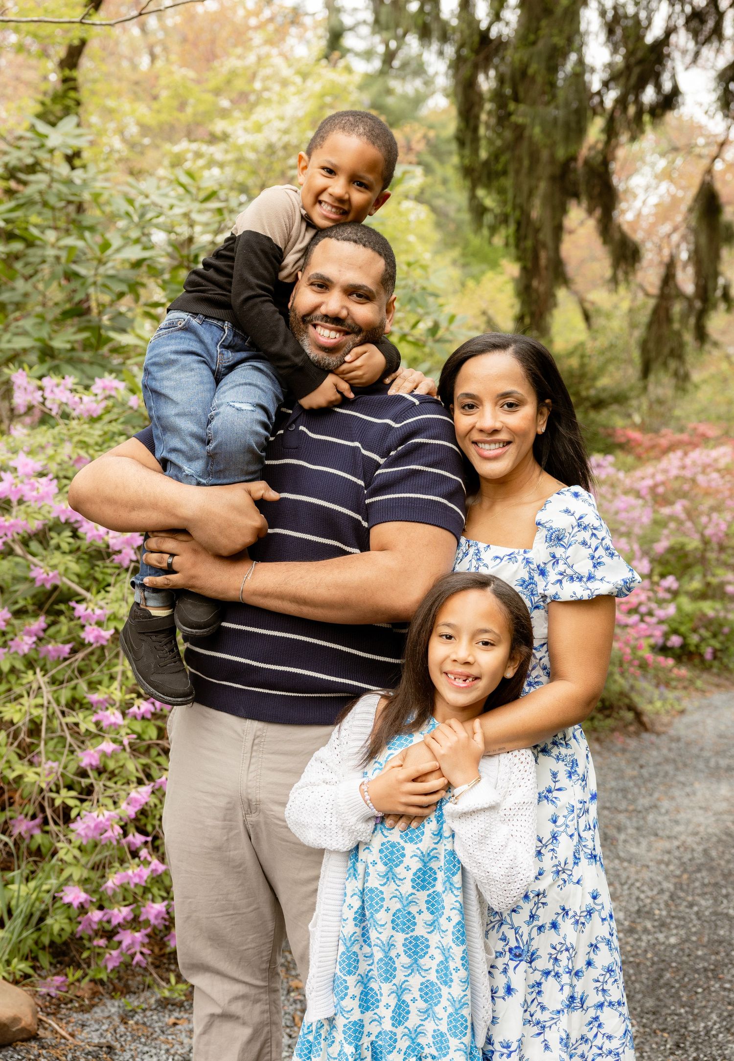 Happy family of four posing outdoors among pink flowering bushes, father holding young son on shoulders with mother and daughter.