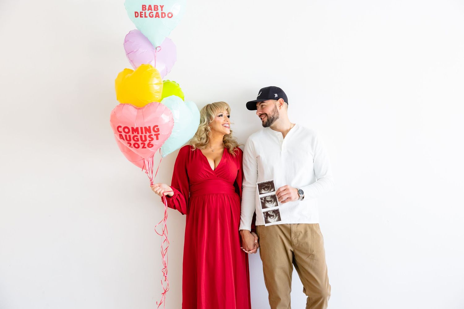 A person in a bright red dress stands next to someone wearing khakis and white shirt against a white background.