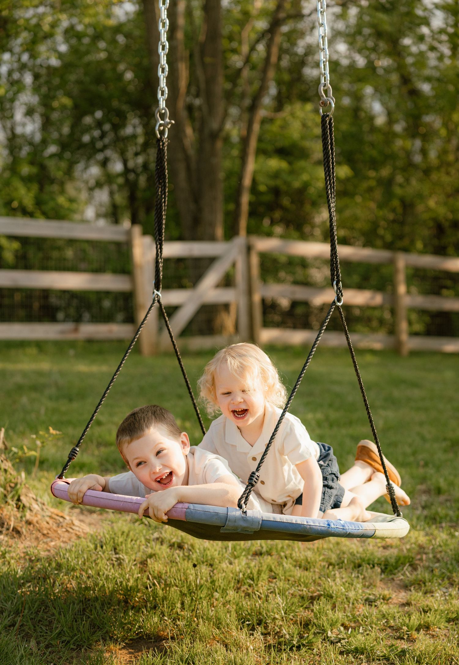 Two happy children laughing together on a large platform swing in a sunny backyard with wooden fence.