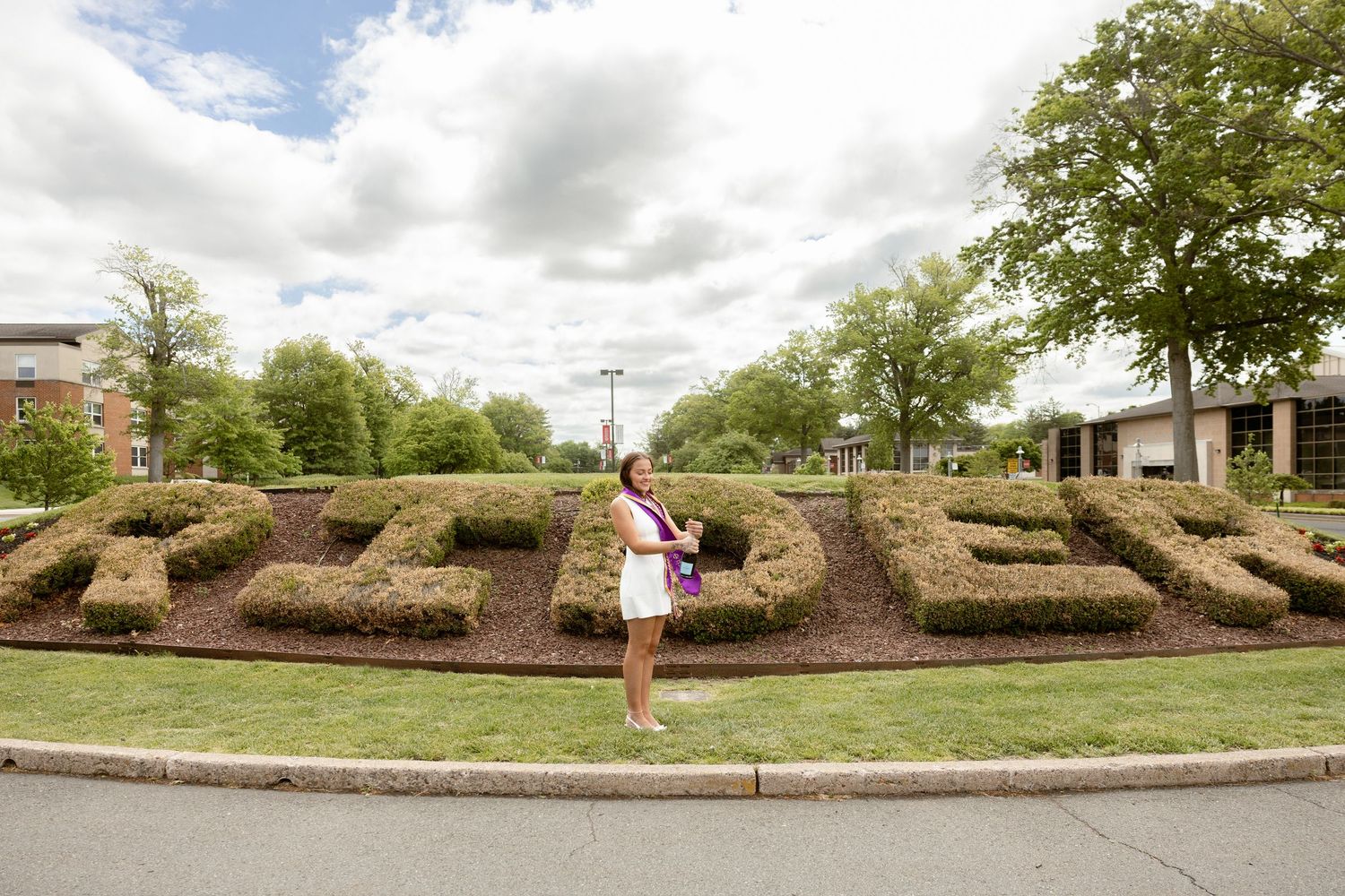 Woman in white dress posing in front of large topiary letters spelling RIDER on a college campus.