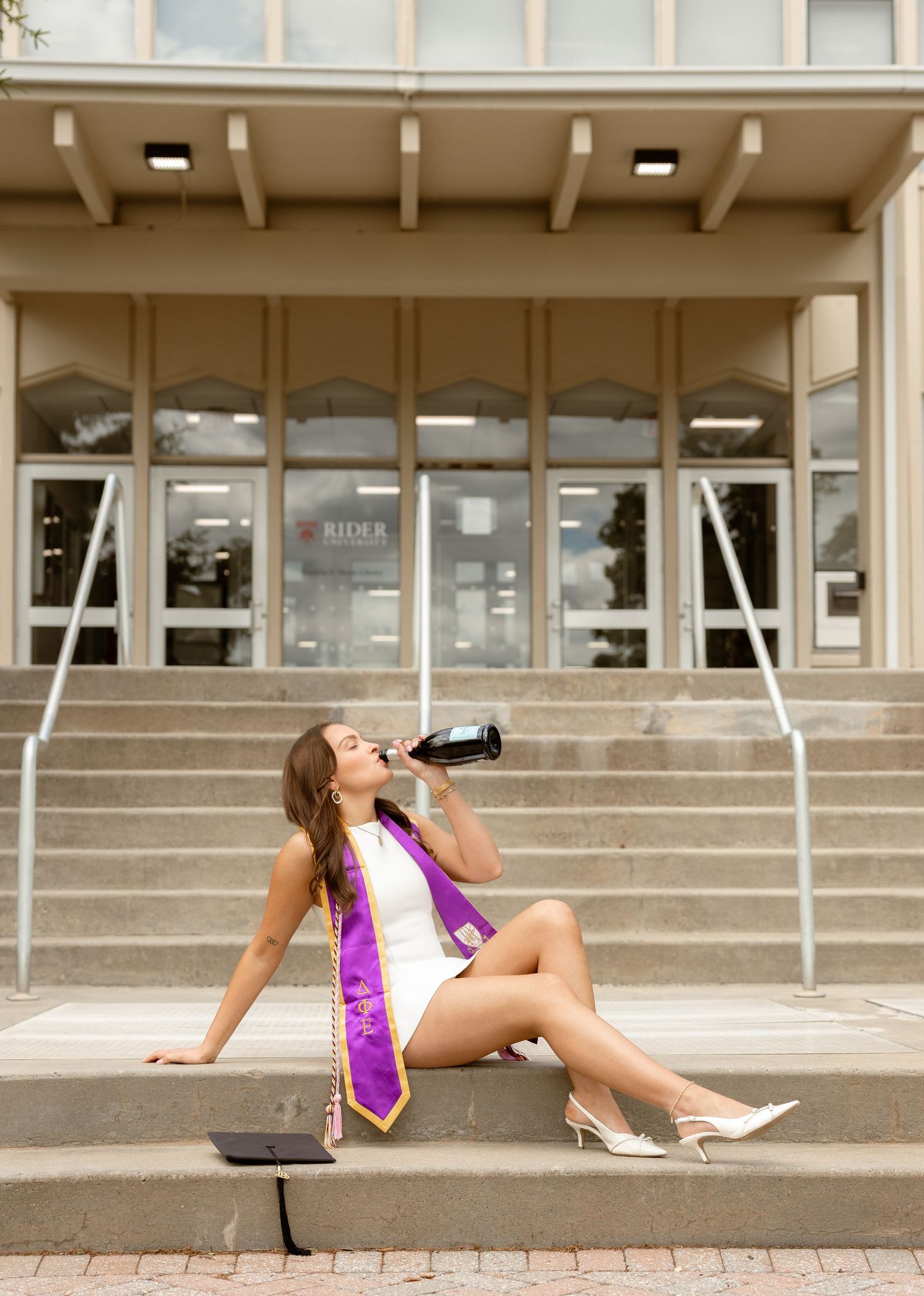 Graduate in white dress and purple stole sits on university steps, drinking from bottle with diploma nearby.
