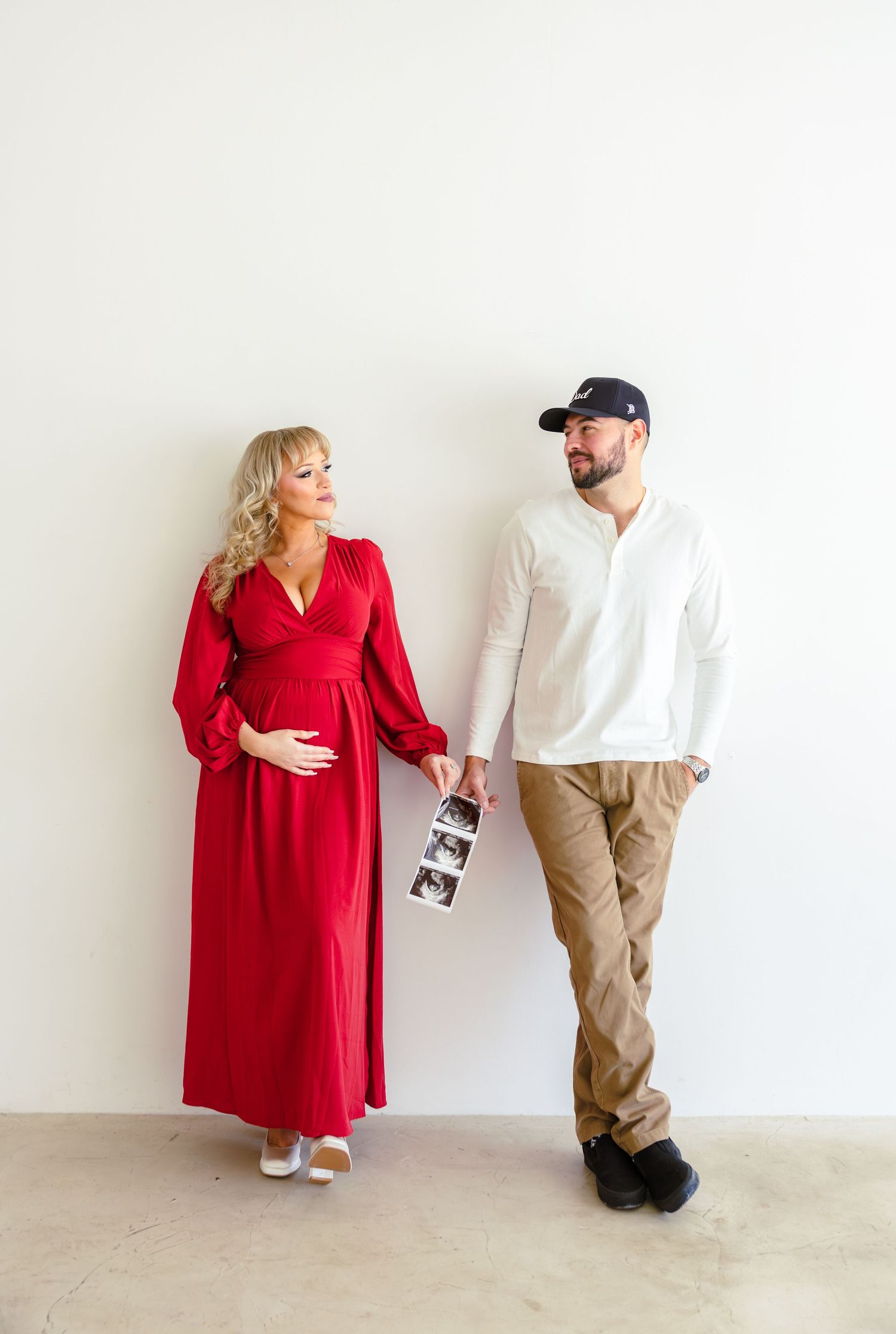 Pregnant woman in red dress and man in white shirt holding ultrasound photos together against white wall.