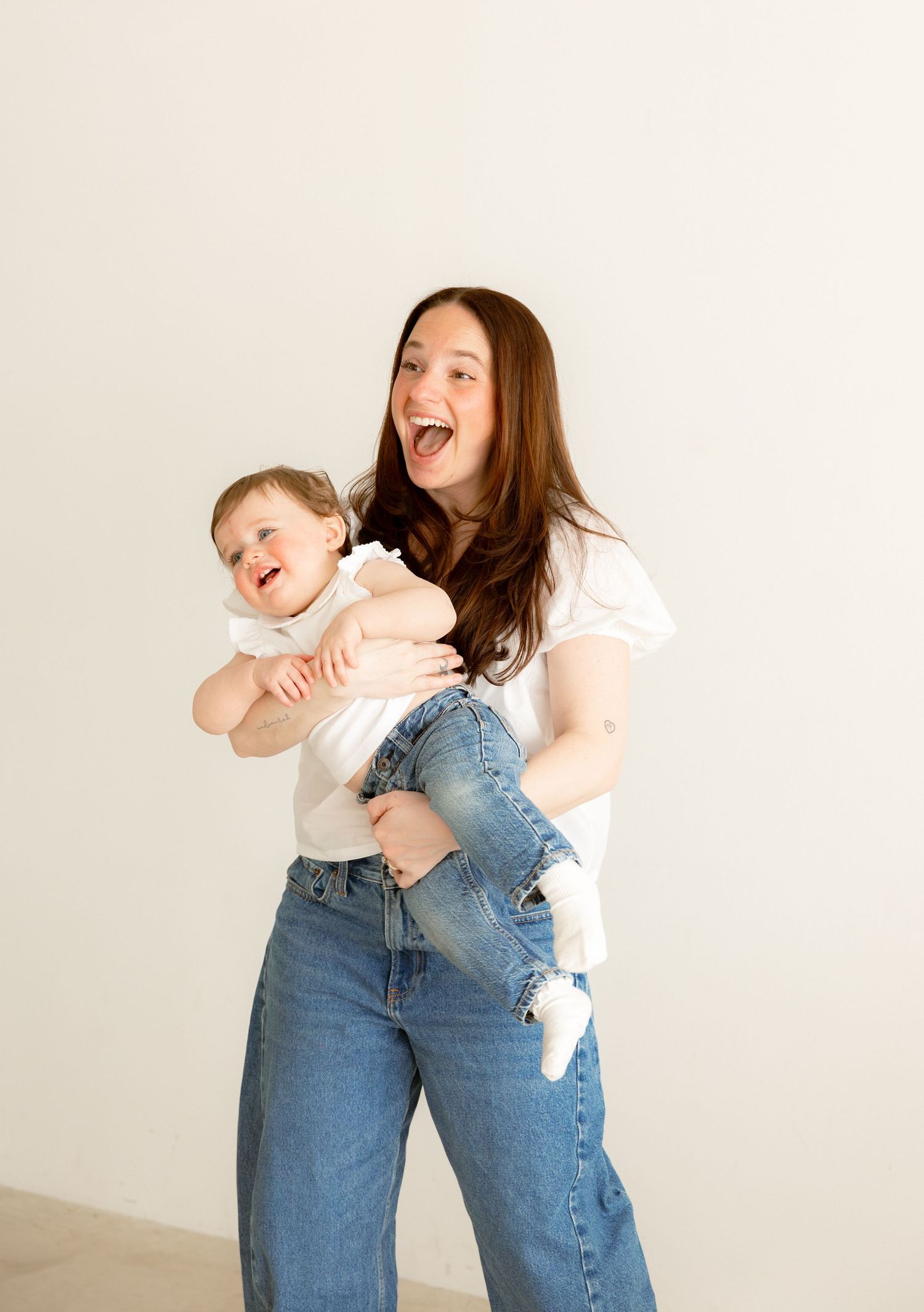 Young woman in white tee and jeans laughing while holding a giggling baby against a white background.
