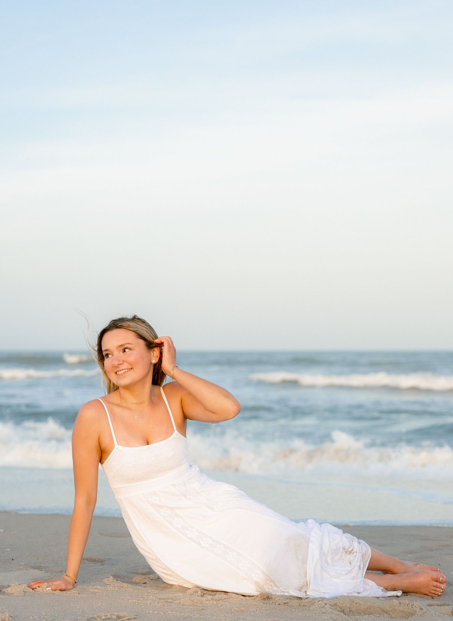 Young woman in white sundress sitting gracefully on sandy beach with ocean waves in background.