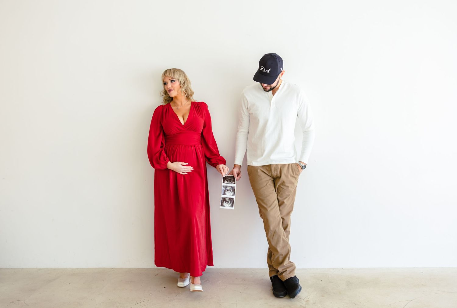 A couple poses against a white wall, with one wearing a flowing red dress and the other in casual attire holding baby shoes.